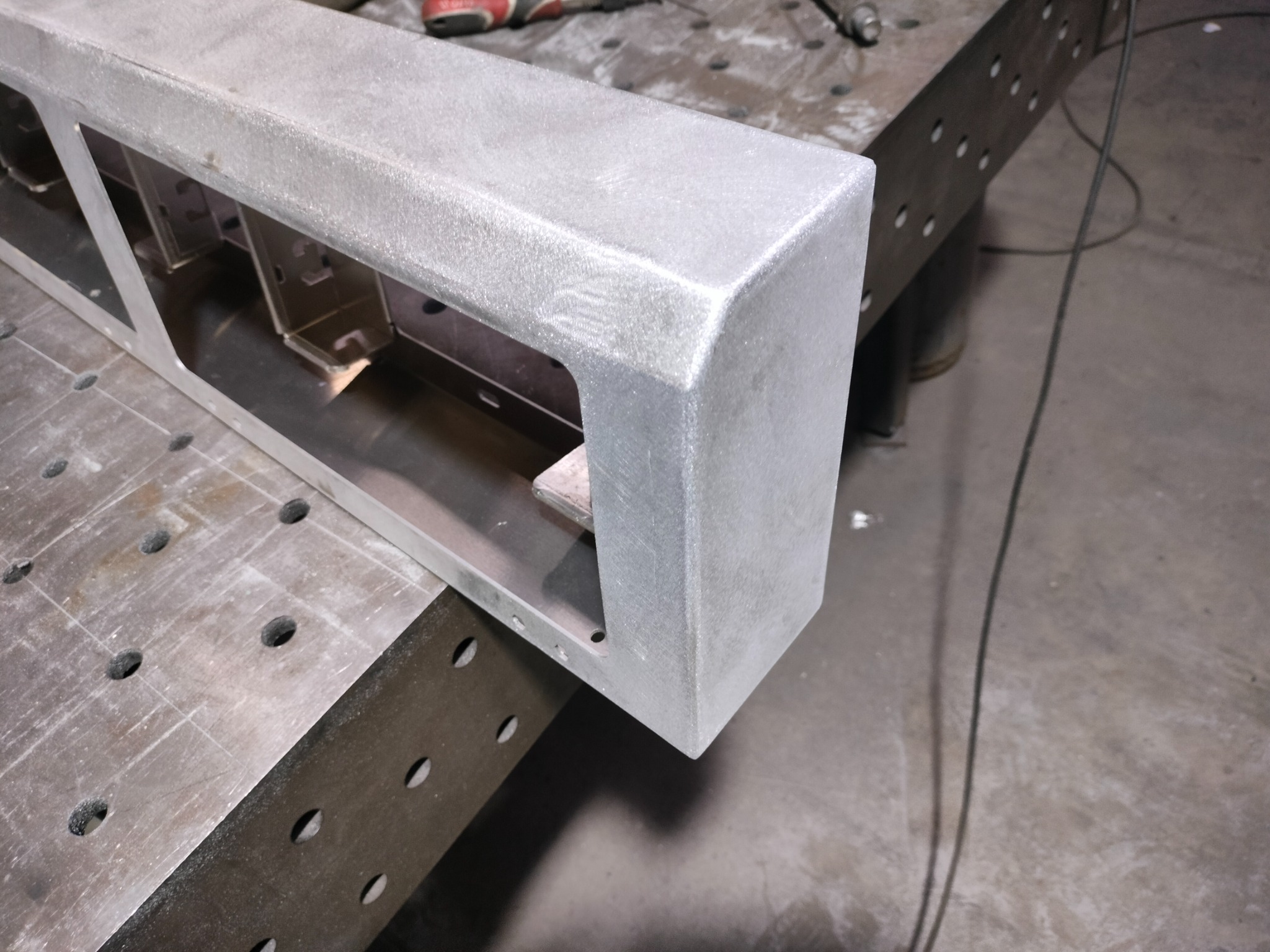 Szlifowana, aluminiowa rama konstrukcyjna z wewnętrznymi wspornikami, leżąca na perforowanym stole warsztatowym, widoczny przewód zasilający i narzędzia w tle.