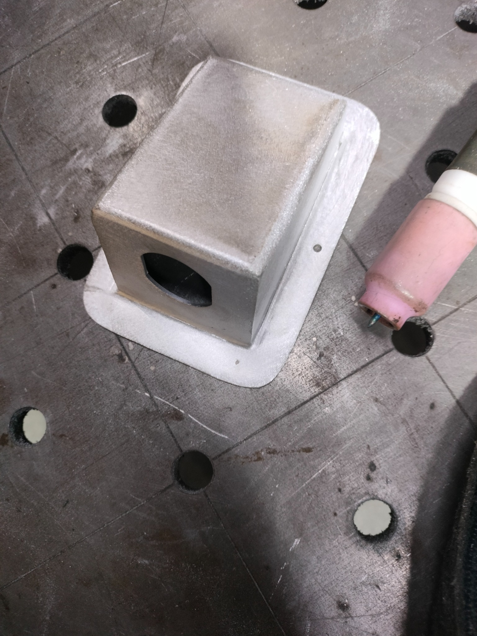 Metalowa obudowa spawana na stole spawalniczym, widoczny uchwyt spawarki TIG, otwór w obudowie.