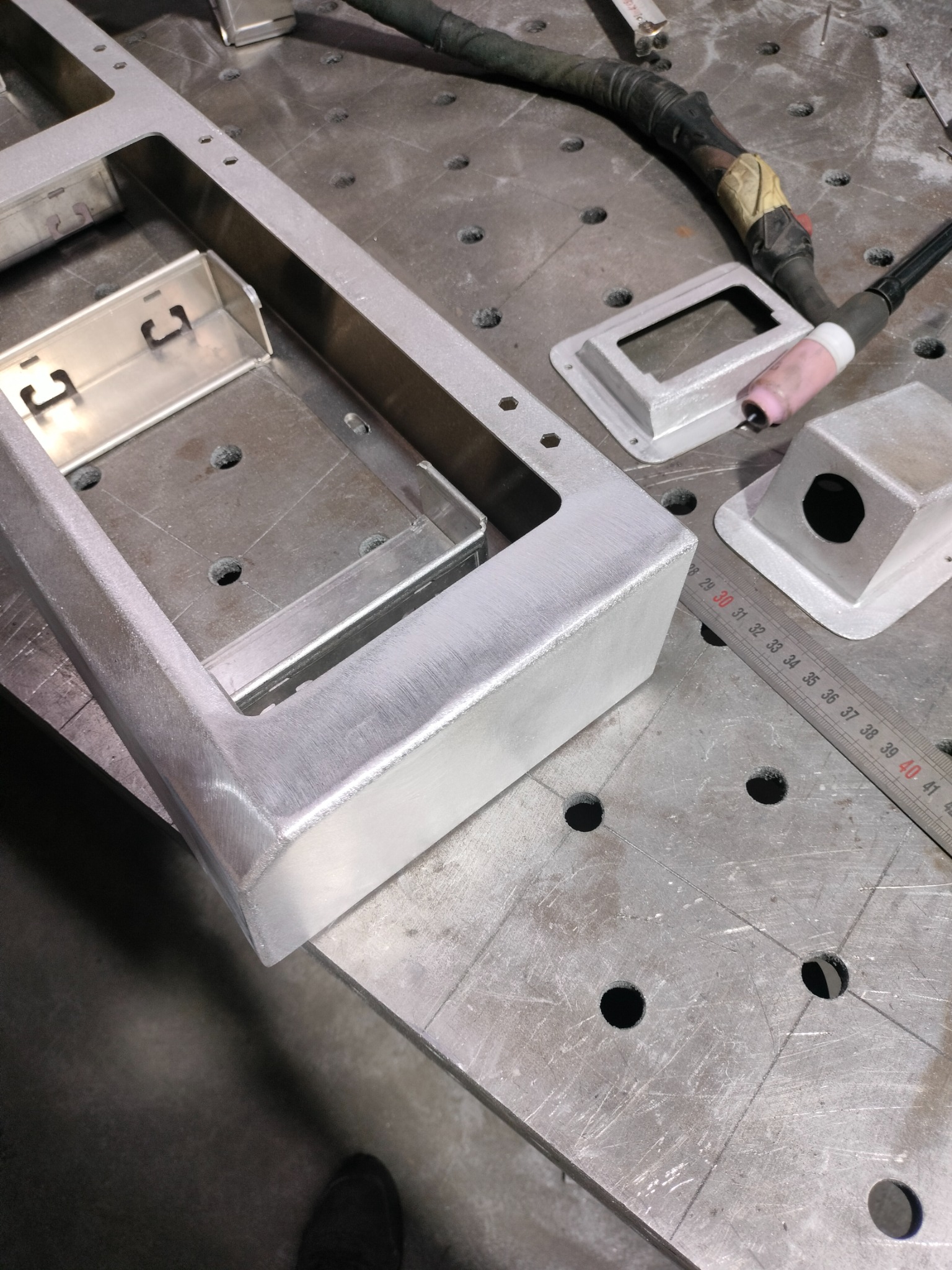 Spawana aluminiowa rama z elementami mocującymi, widoczna na stole spawalniczym z narzędziami i miarką.