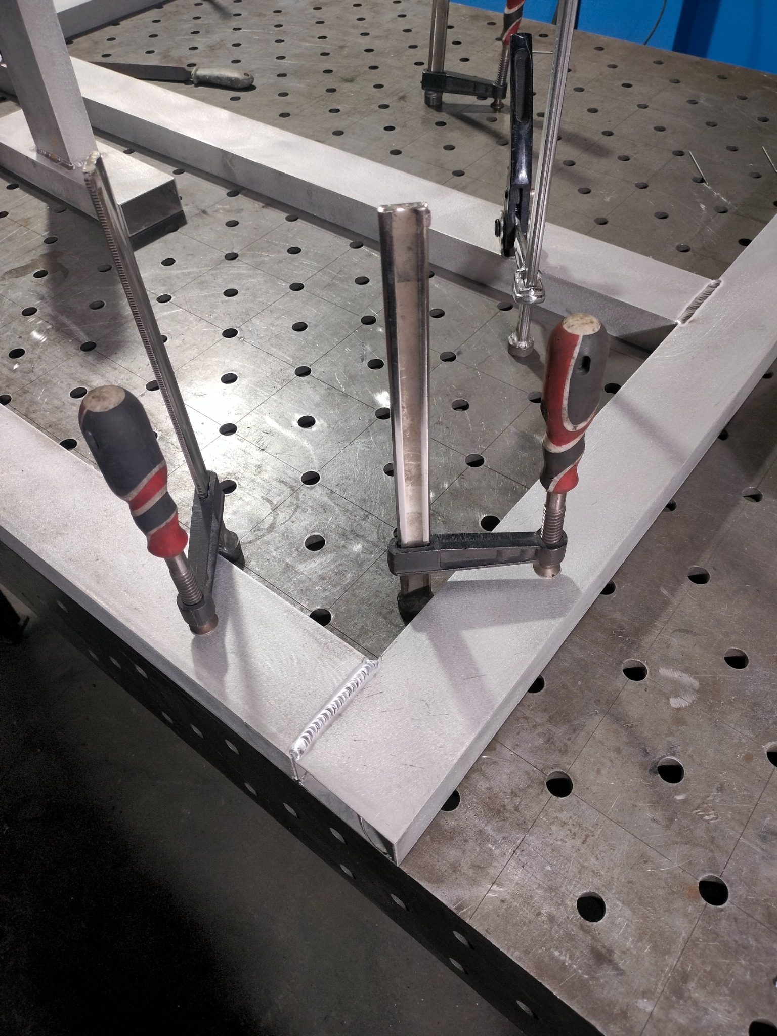 Spawana rama z profili aluminiowych, zabezpieczona zaciskami na perforowanym stole spawalniczym, widoczne spoiny.