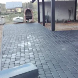 Ułożona, szara kostka brukowa na podwórku z widocznym grillem z cegły, paletami z materiałami budowlanymi i młodymi tujami w tle.