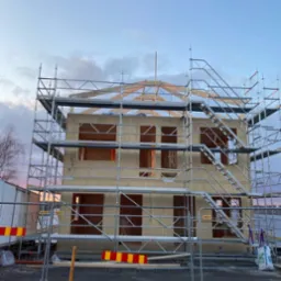 Surowy stan dwukondygnacyjnego domu w konstrukcji szkieletowej drewnianej, otoczonego rusztowaniami i tymczasowym ogrodzeniem budowlanym, widoczne elementy konstrukcyjne dachu i okien.