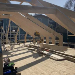 Konstrukcja drewnianego dachu w trakcie budowy, widoczne krokwie, płatwie, oraz piła elektryczna na stojaku, w tle budynki mieszkalne.