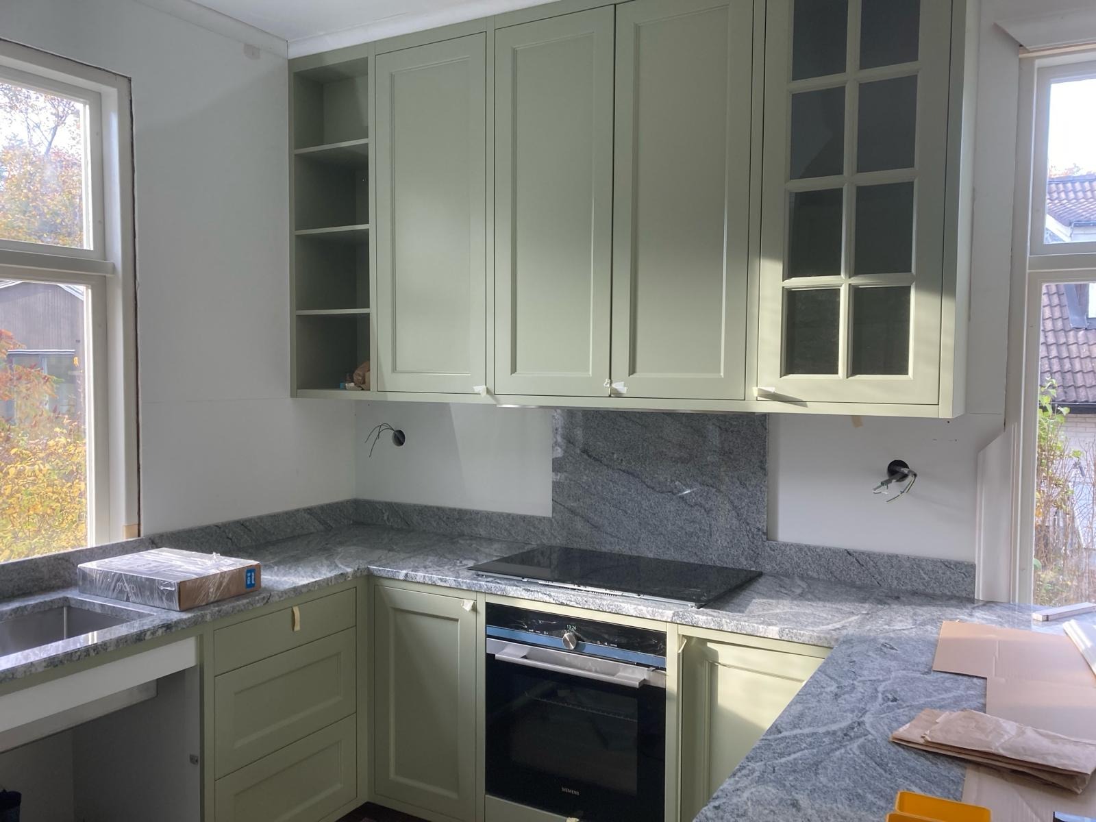 Kuchnia w trakcie montażu: zielone szafki wiszące i dolne, szary granitowy blat, płyta indukcyjna i piekarnik w zabudowie, widoczne niedokończone instalacje elektryczne.