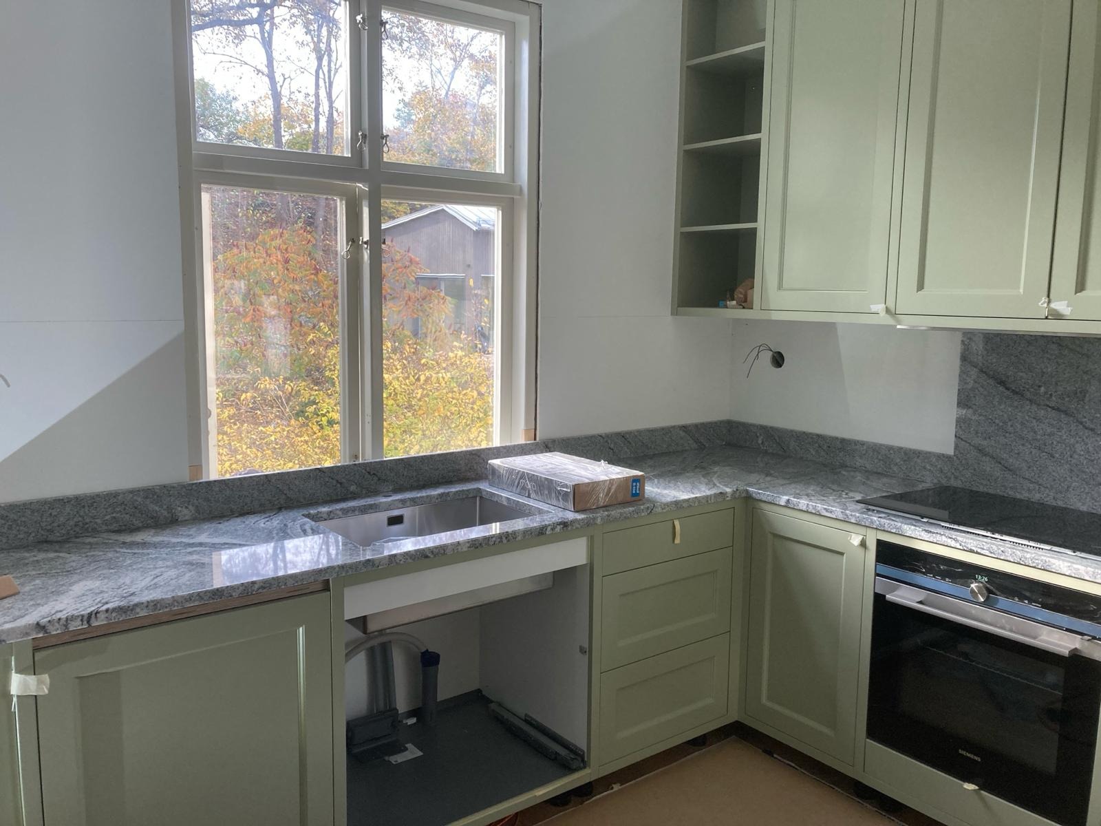 Wnętrze kuchni w trakcie wykańczania: zielone szafki, szary granitowy blat, stalowy zlew z zapakowanym syfonem, okno z widokiem na jesienne drzewa, płyta indukcyjna i piekarnik Siemens.