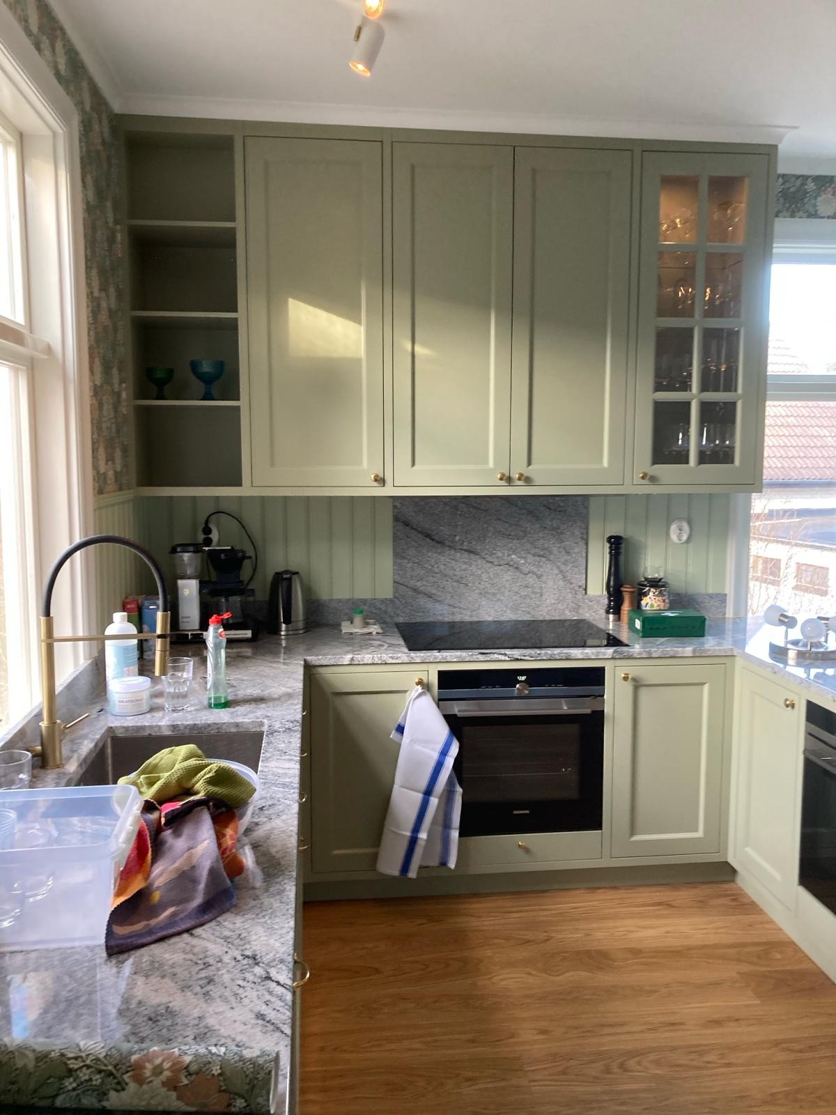 Jasnozielona kuchnia z granitowym blatem i złotą armaturą, widoczne szafki wiszące i sprzęt AGD, w zlewie naczynia i ściereczki.