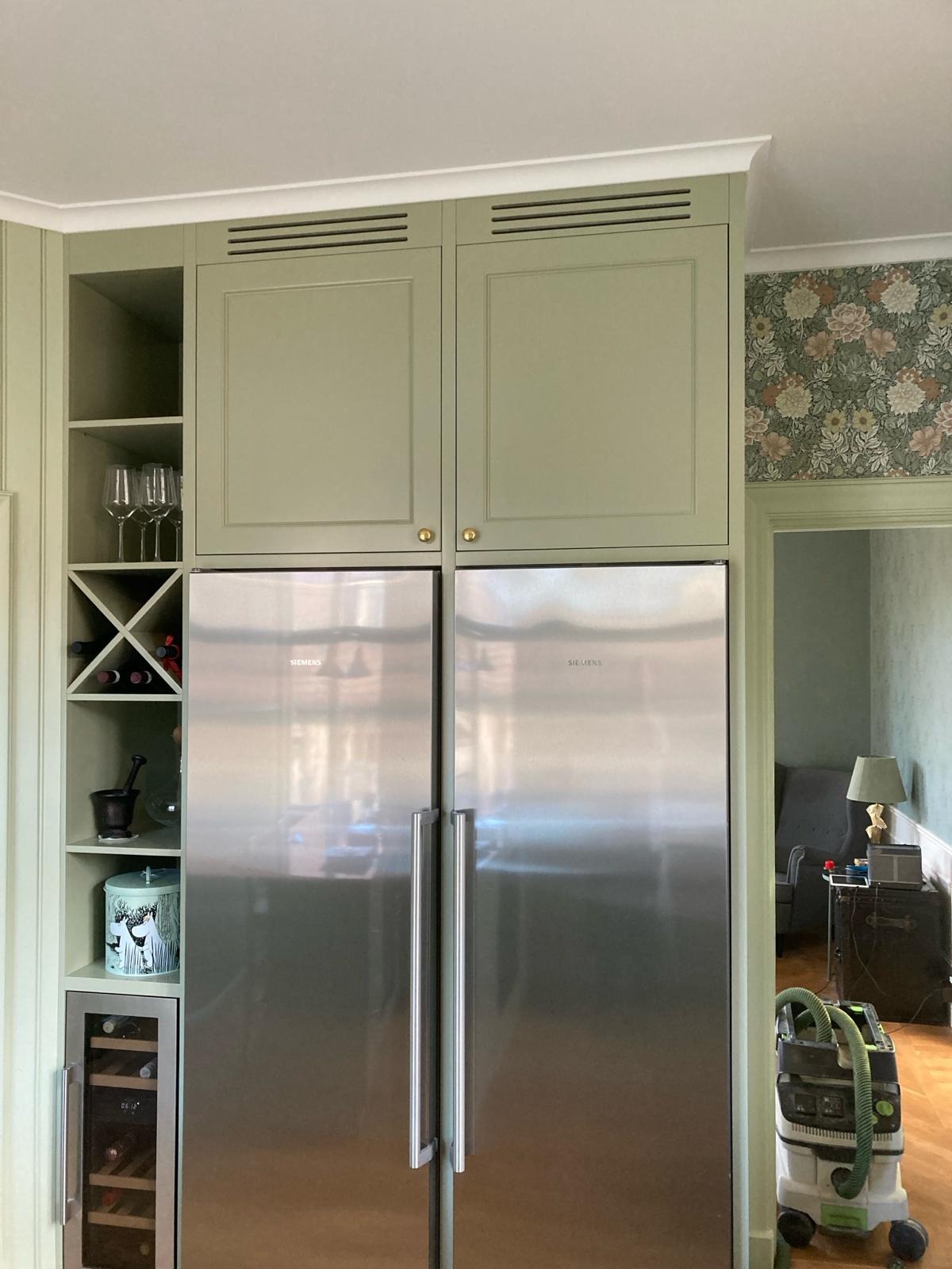 Kuchnia w stylu retro: zabudowa meblowa w odcieniach zieleni z lodówką side-by-side Siemens, półkami na wino i dekoracyjną tapetą w kwiaty w tle, widoczny odkurzacz przemysłowy.