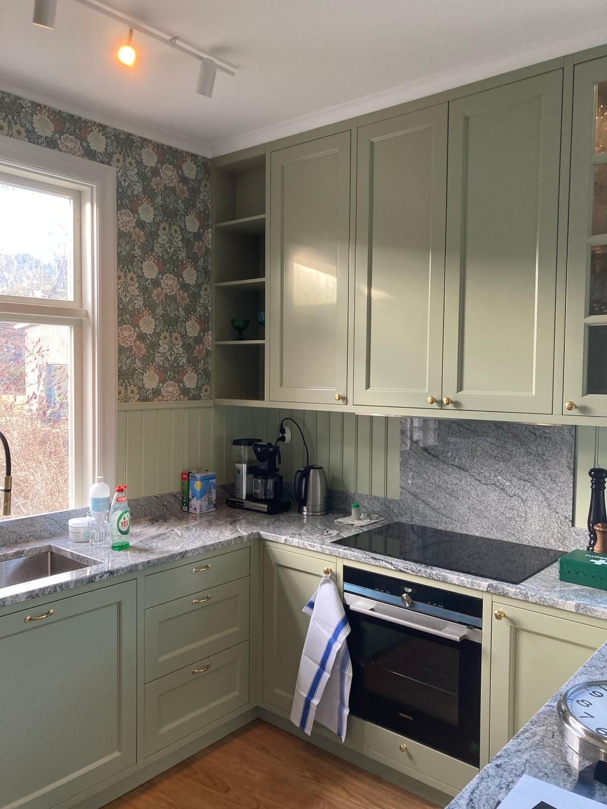 Jasnozielona kuchnia z granitowym blatem, wzorzystą tapetą i nowoczesną płytą indukcyjną.