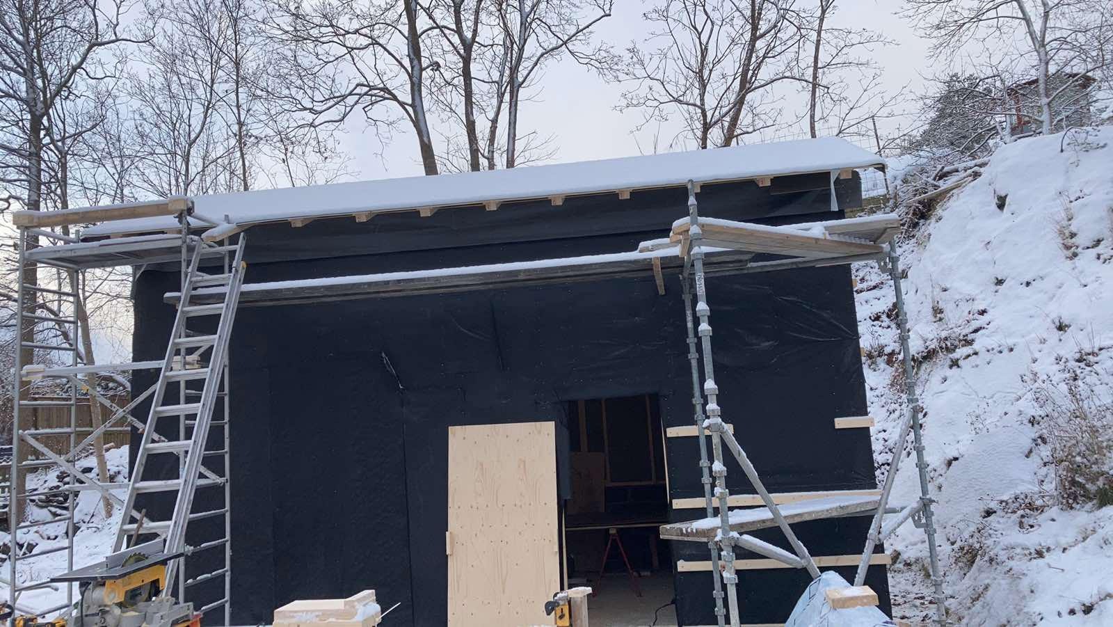 Budynek w trakcie ocieplania, pokryty czarną folią, widoczne rusztowania i śnieg na dachu oraz ziemi.