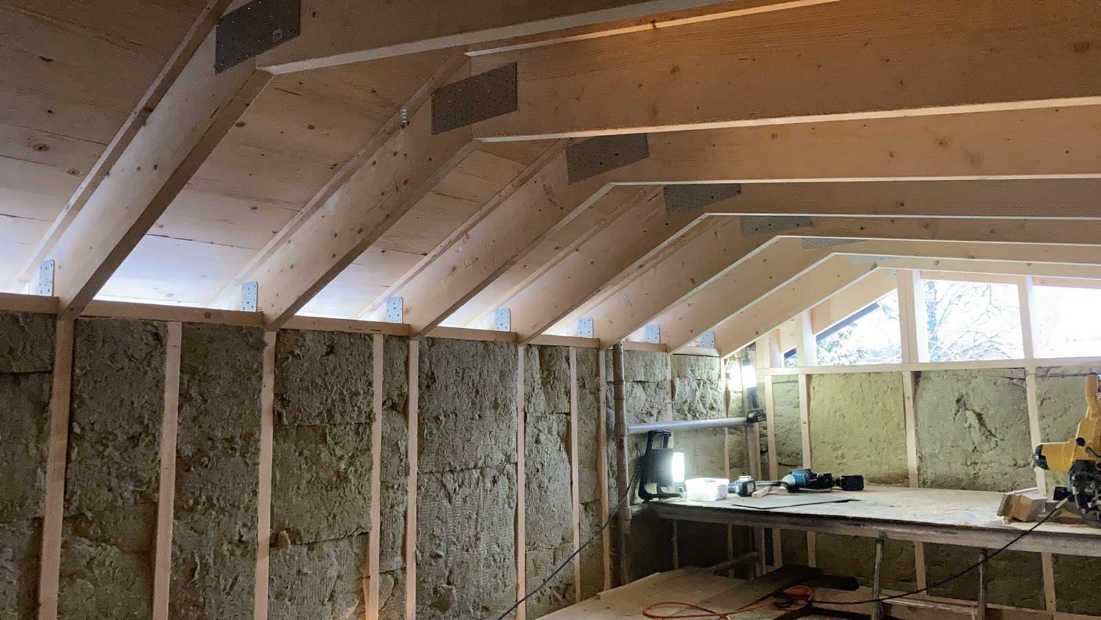Poddasze w trakcie ocieplania wełną mineralną, widoczne drewniane krokwie z metalowymi łącznikami, narzędzia na stole roboczym i okno z widokiem na drzewa.