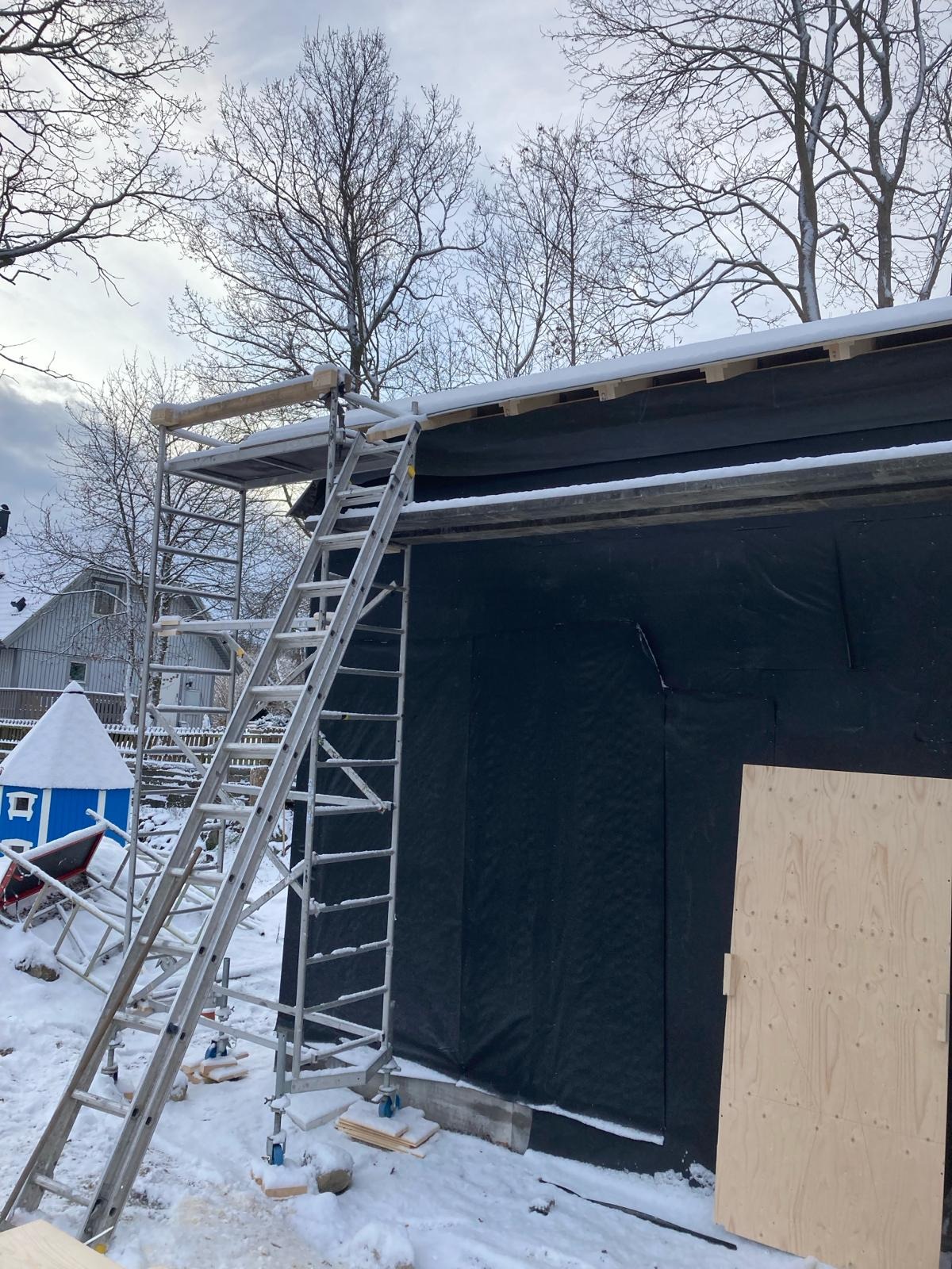 Elewacja budynku w trakcie ocieplania, zabezpieczona czarną folią, z rusztowaniem i drabiną w zimowej scenerii.