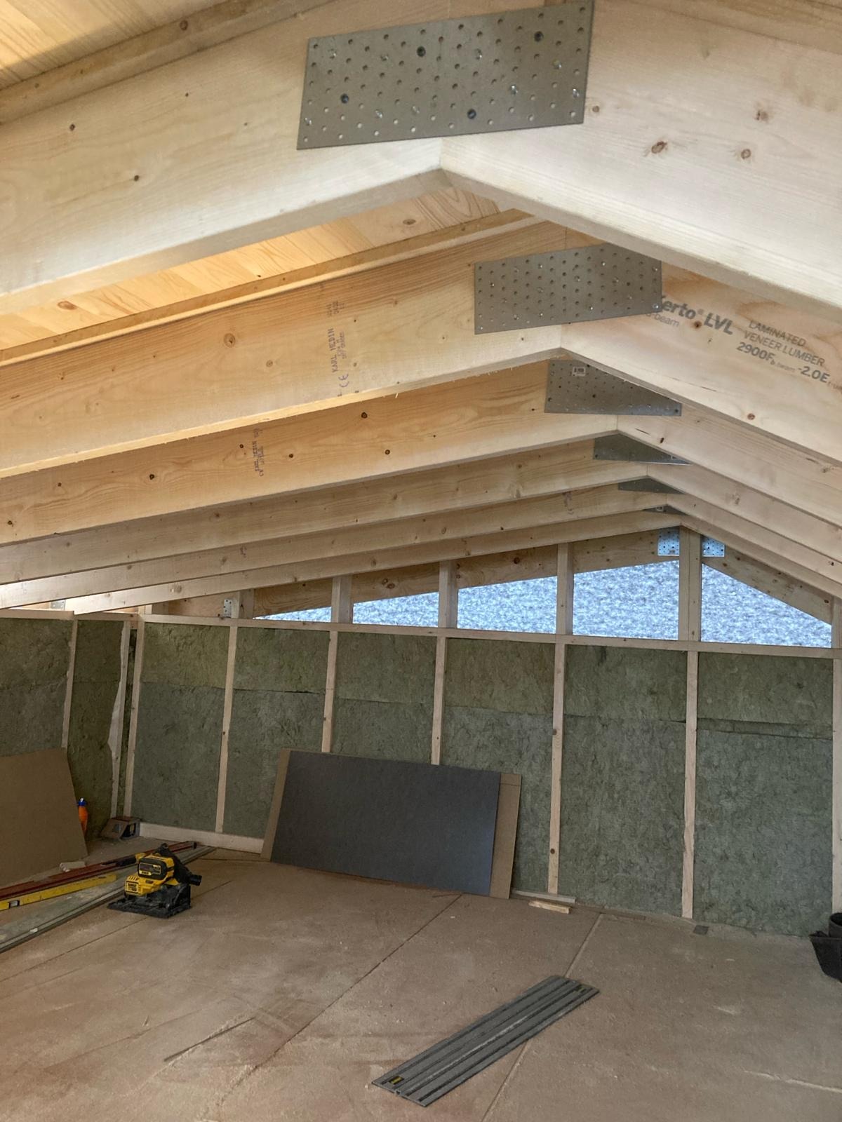 Wnętrze poddasza w trakcie ocieplania: widoczne drewniane belki stropowe z metalowymi łącznikami, ściany wypełnione materiałem izolacyjnym, płyty kartonowo-gipsowe oparte o ścianę, narzędzia...