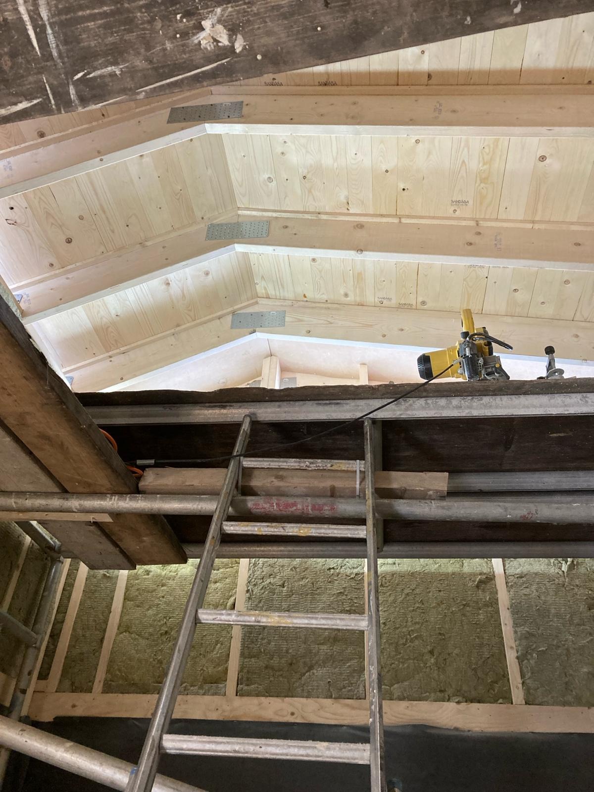 Szkielet drewnianej konstrukcji poddasza z widoczną izolacją z wełny mineralnej między krokwiami, rusztowanie z drabiną oraz narzędzia na górze.