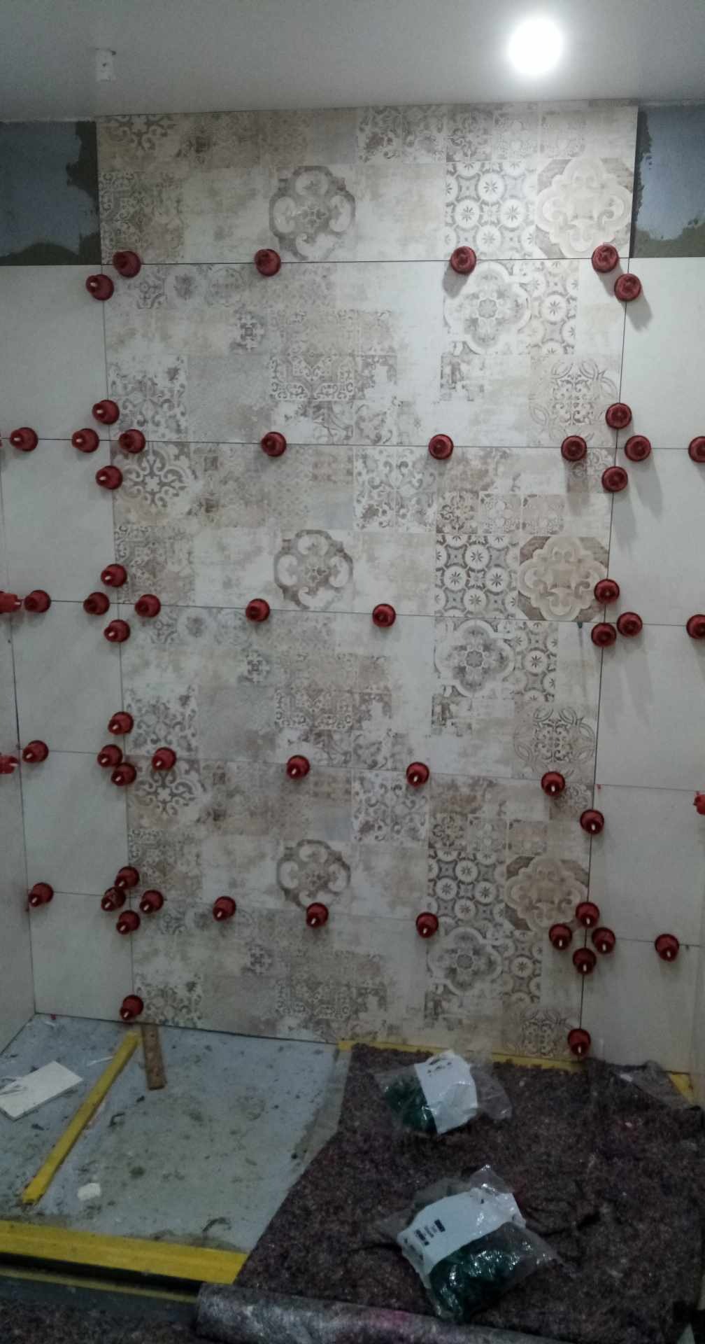 Ściana w trakcie układania płytek z wzorem, z widocznymi czerwonymi klipsami poziomującymi i zapasem płytek na podłodze.