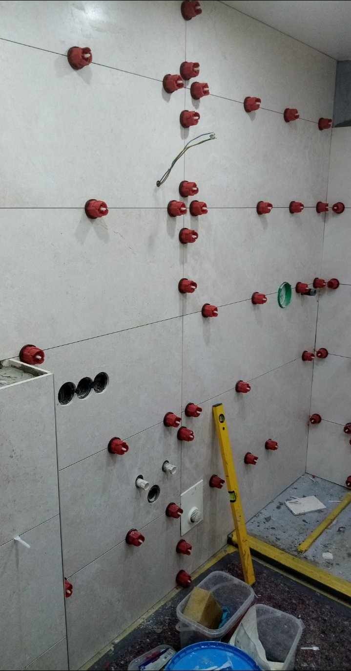 Świeżo ułożone, jasnoszare płytki ceramiczne na ścianie łazienki, zabezpieczone czerwonymi klipsami poziomującymi, widoczne otwory na instalacje i żółta poziomica.