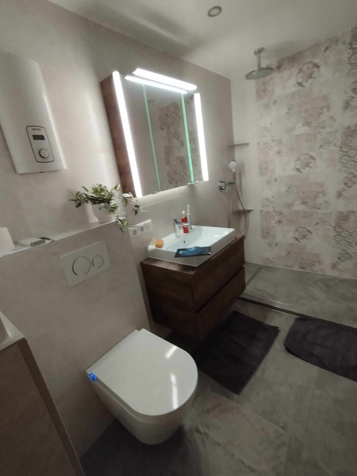 Nowoczesna łazienka z podwieszaną toaletą, drewnianą szafką pod umywalką z podświetlanym lustrem i kabiną prysznicową z dekoracyjnymi płytkami.