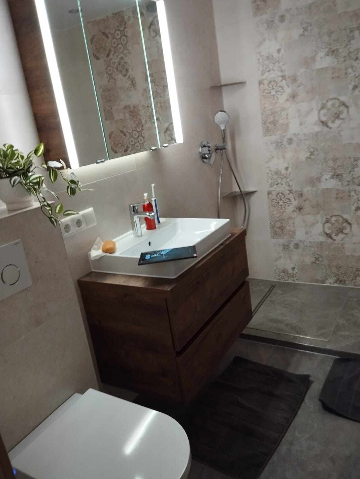 Nowoczesna łazienka z umywalką nablatową na szafce z ciemnego drewna, wzorzyste płytki na ścianie, podświetlane lustro, i widocznym odpływem liniowym w podłodze kabiny prysznicowej.