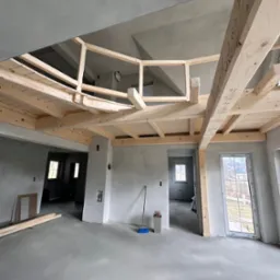 Surowa antresola z jasnego drewna w szarym wnętrzu. Widać drewniane elementy konstrukcyjne i surowy beton na podłodze. W tle okna i drzwi.