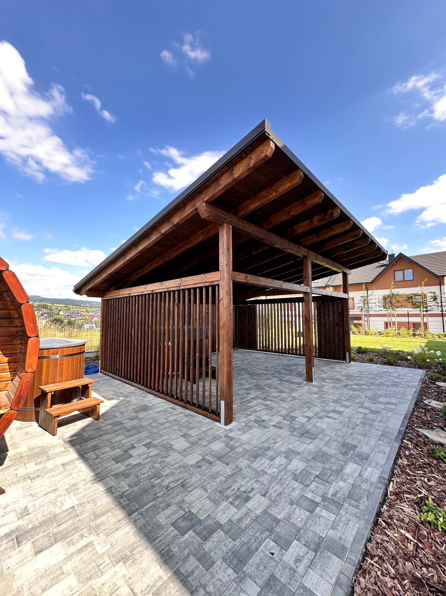 Drewniana altana z ażurowymi ścianami i płaskim dachem, na szarym tarasie z kostki. Obok drewniana beczka z hydromasażem. Błękitne niebo w tle.