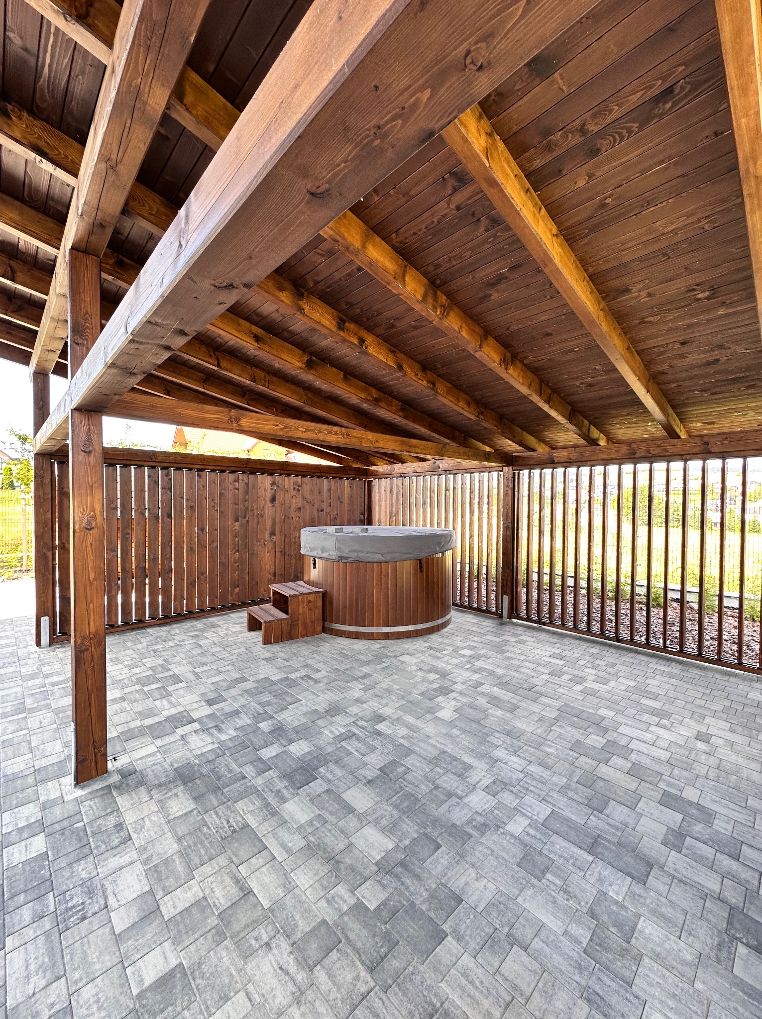 Drewniana altana z jacuzzi na szarym brukowanym tarasie. Widoczne drewniane słupy, zadaszenie i ażurowe ścianki. Całość utrzymana w ciepłej kolorystyce.