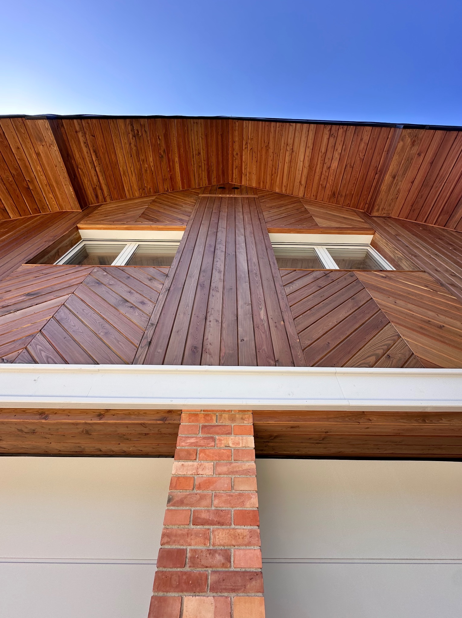 Drewniana elewacja domu z unikalnym wzorem ułożenia desek, widok z dołu, podkreślony ceglany filar i białe okna na tle błękitnego nieba.
