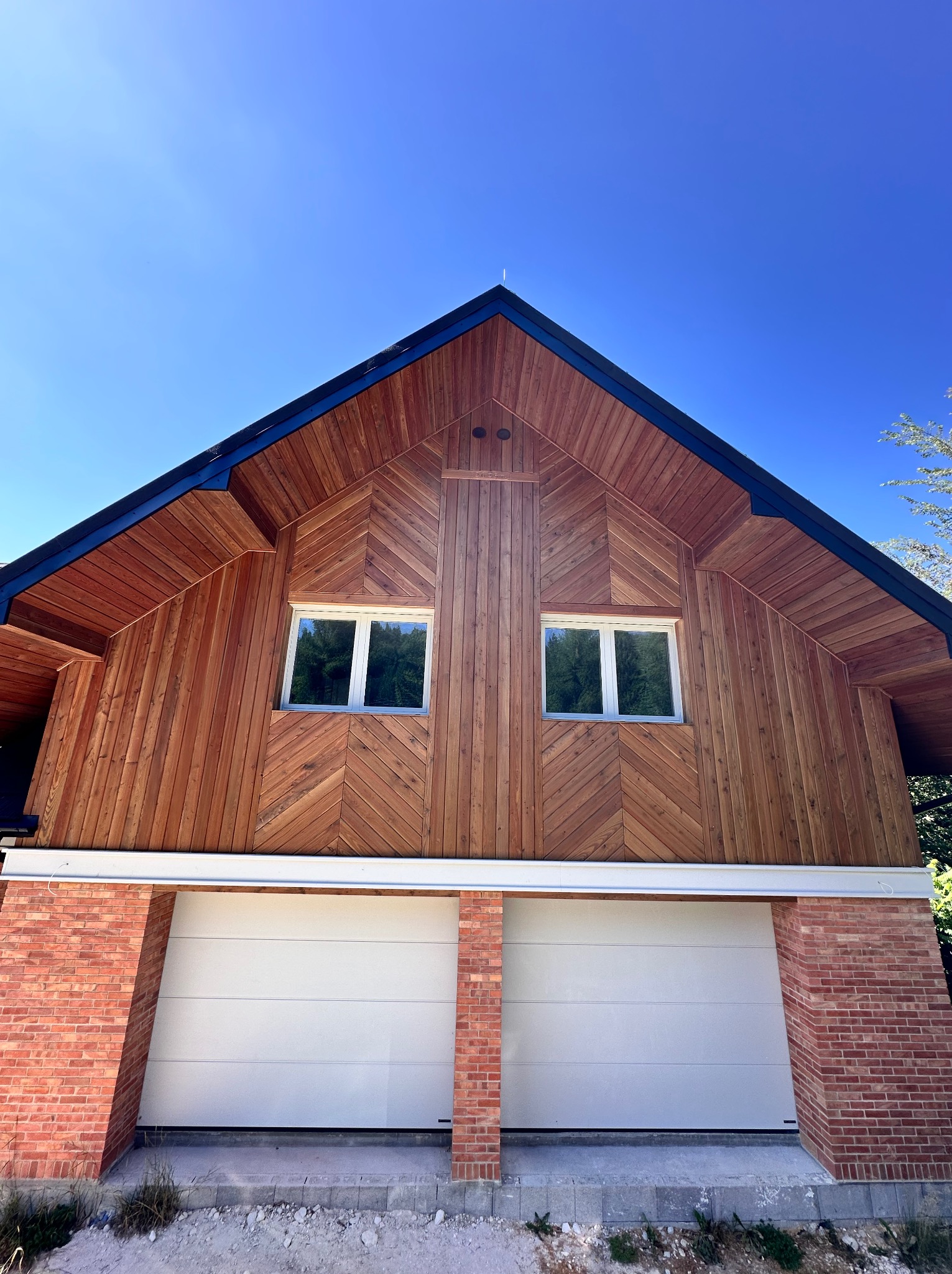 Wykończona elewacja budynku z drewnianą okładziną ułożoną we wzór jodełki, dwa okna i dwustanowiskowy garaż z ceglaną podstawą w słoneczny dzień.