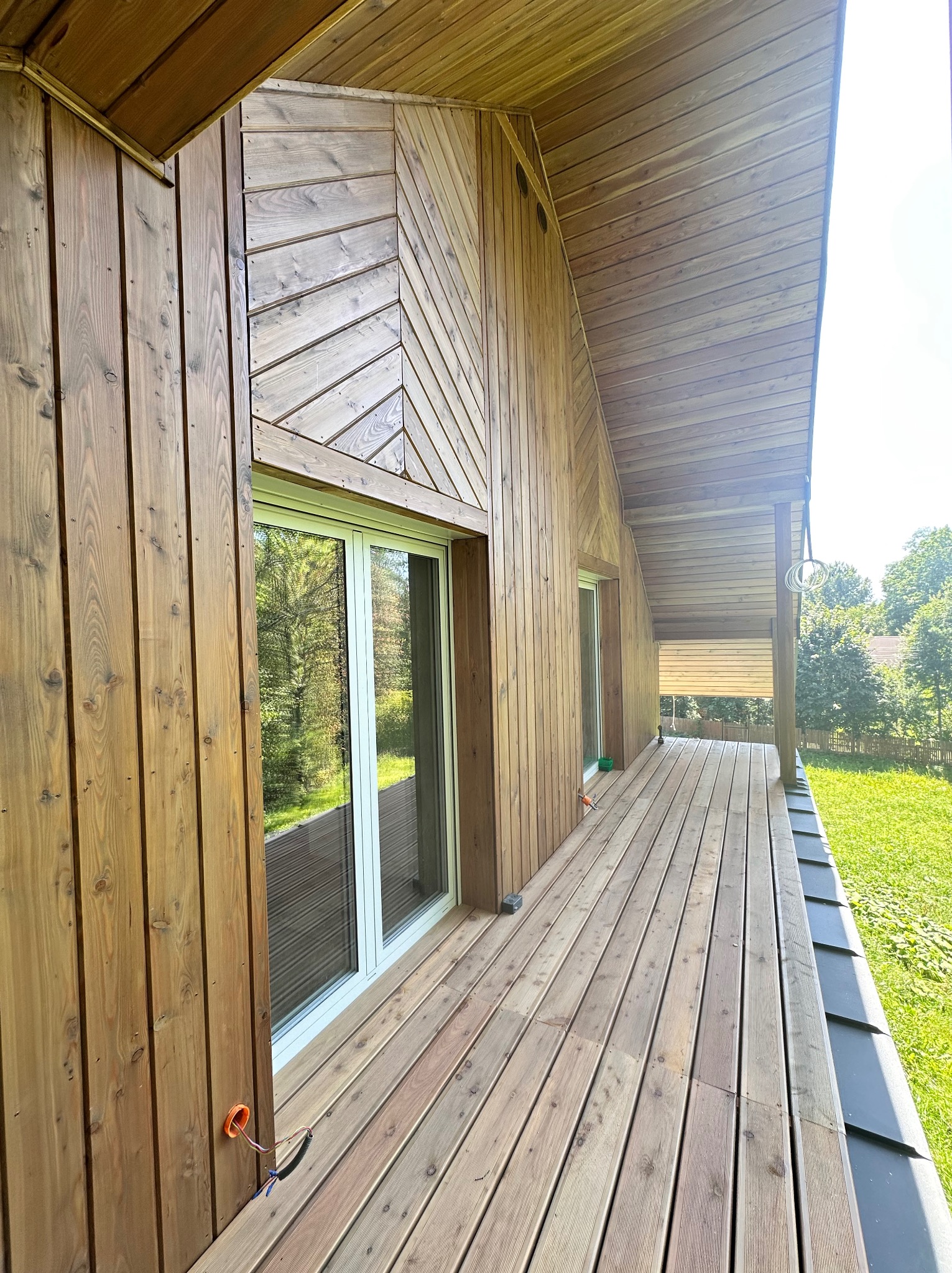 Drewniana elewacja domu z tarasem. Widoczne deski ułożone w różnych kierunkach, okno z widokiem na zieleń i fragment instalacji elektrycznej na zewnątrz budynku.