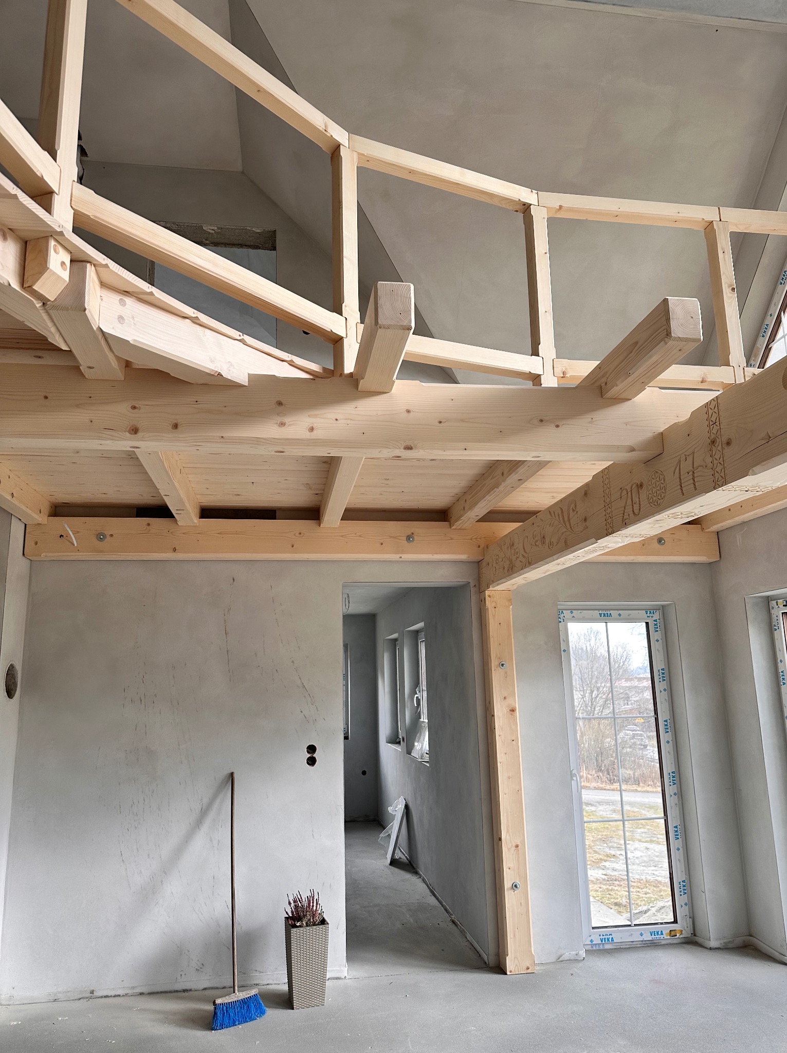 Surowe drewno antresoli w trakcie montażu w jasnym wnętrzu z oknami. Na podłodze leży miotła i stoi doniczka z kwiatami. Widoczne zdobienia na belkach.
