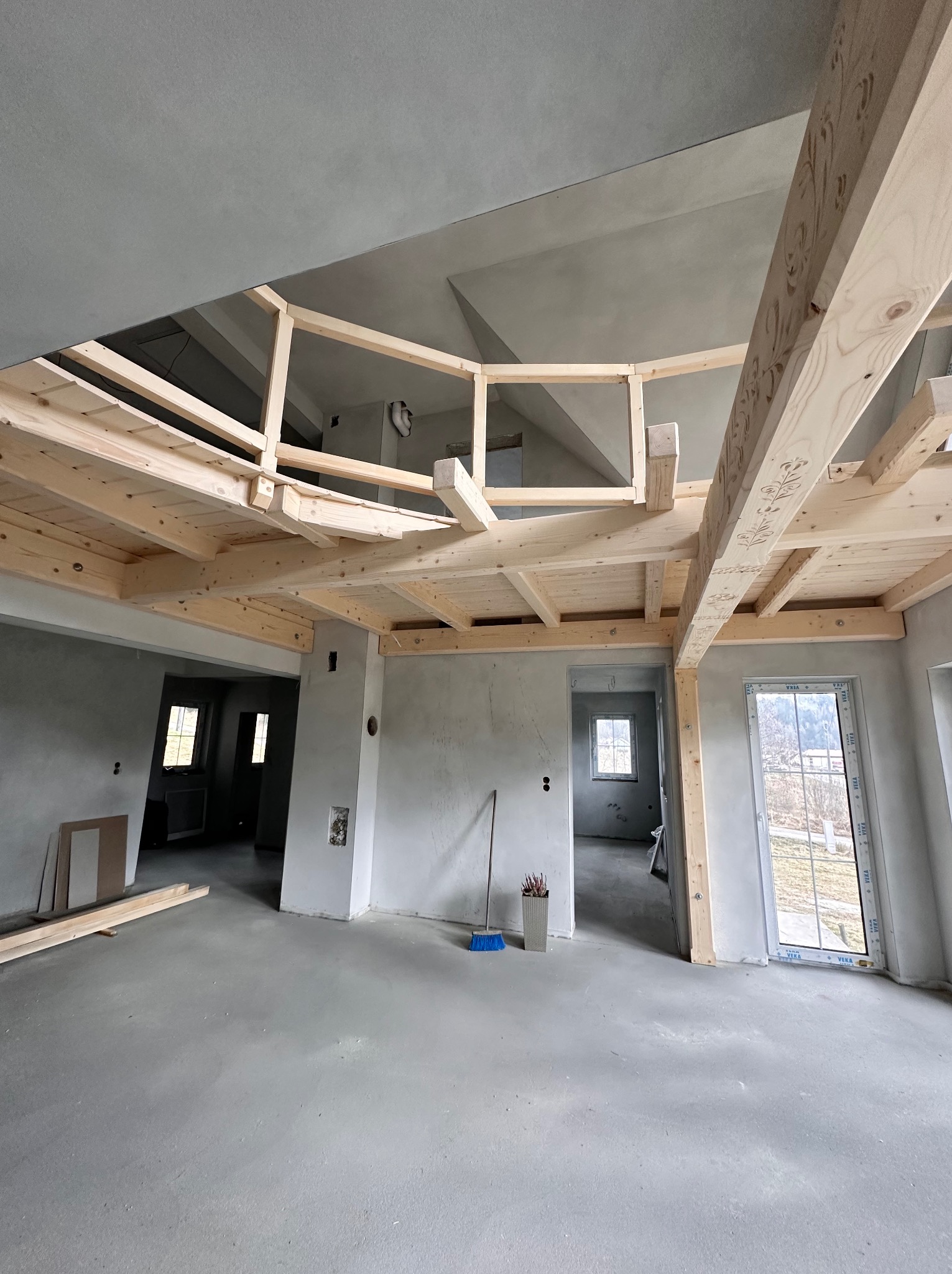 Surowa antresola z jasnego drewna w szarym wnętrzu. Widać drewniane elementy konstrukcyjne i surowy beton na podłodze. W tle okna i drzwi.