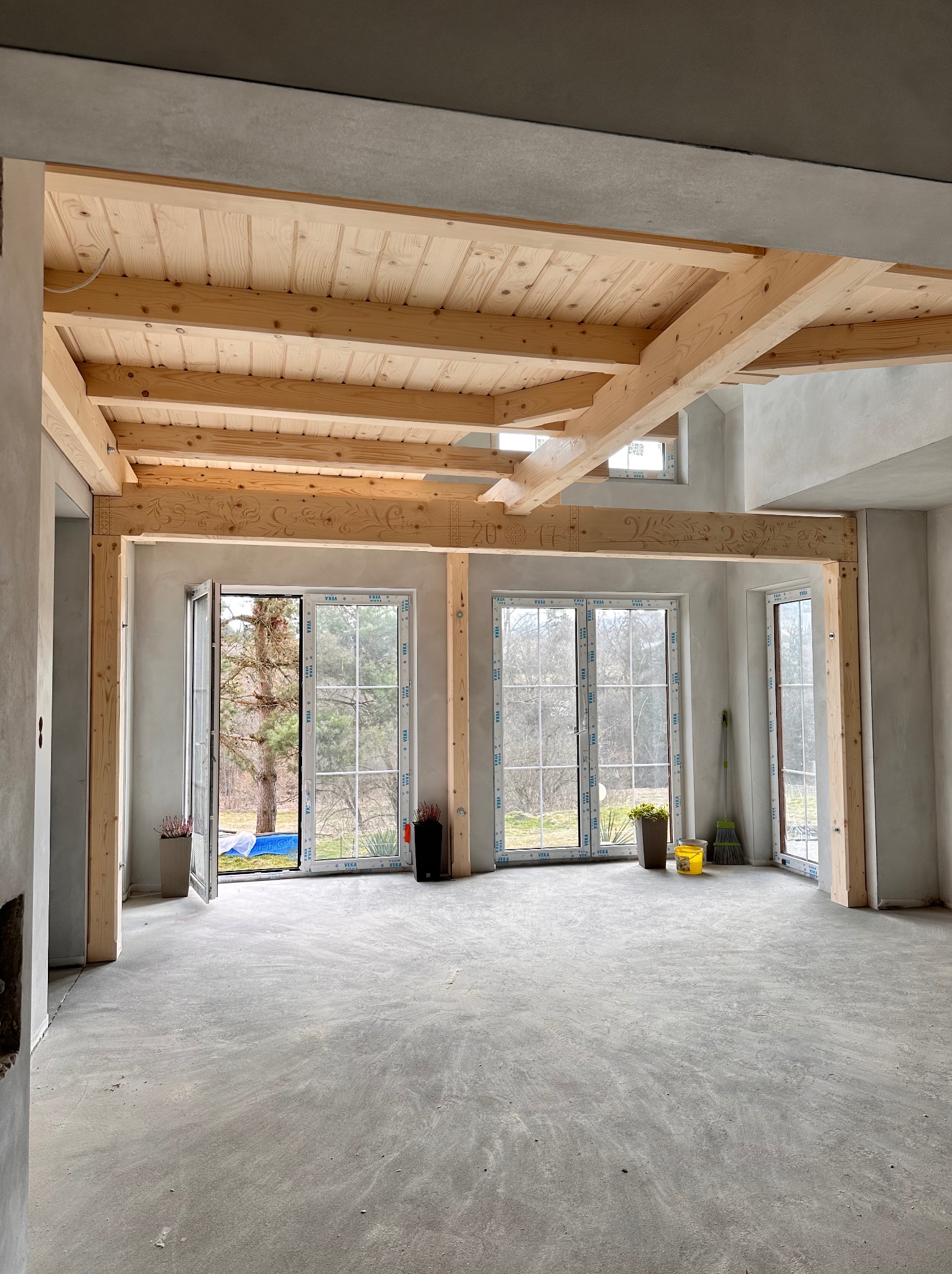 Surowy stan wnętrza z drewnianą konstrukcją stropu i ozdobnymi belkami. Widoczne okna z widokiem na zieleń, surowa podłoga i ściany w odcieniach szarości.