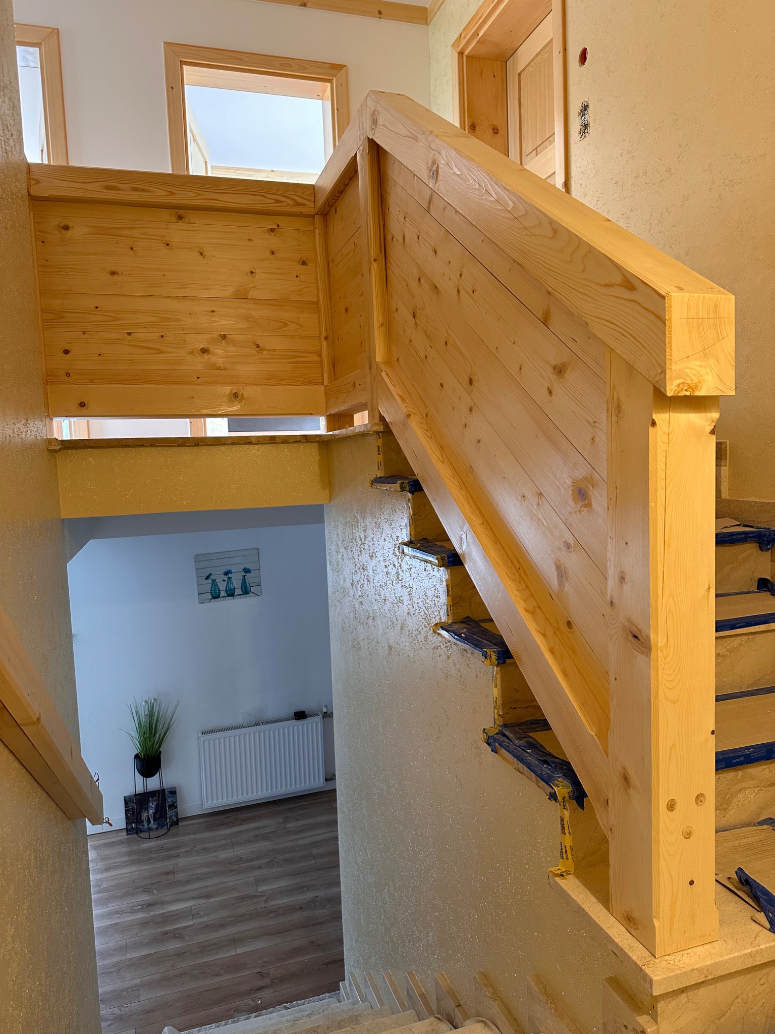Jasne, drewniane schody z balustradą w trakcie montażu. Widoczne zabezpieczenia na stopniach. W tle widoczne pomieszczenie z grzejnikiem i dekoracją.
