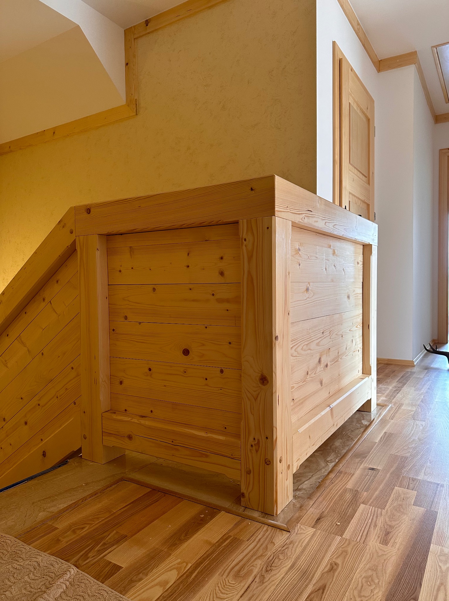 Drewniana zabudowa schodów z surowego drewna, widoczna z boku, z elementami wykończeniowymi w tle. Naturalne oświetlenie podkreśla strukturę drewna i ciepłą kolorystykę wnętrza.