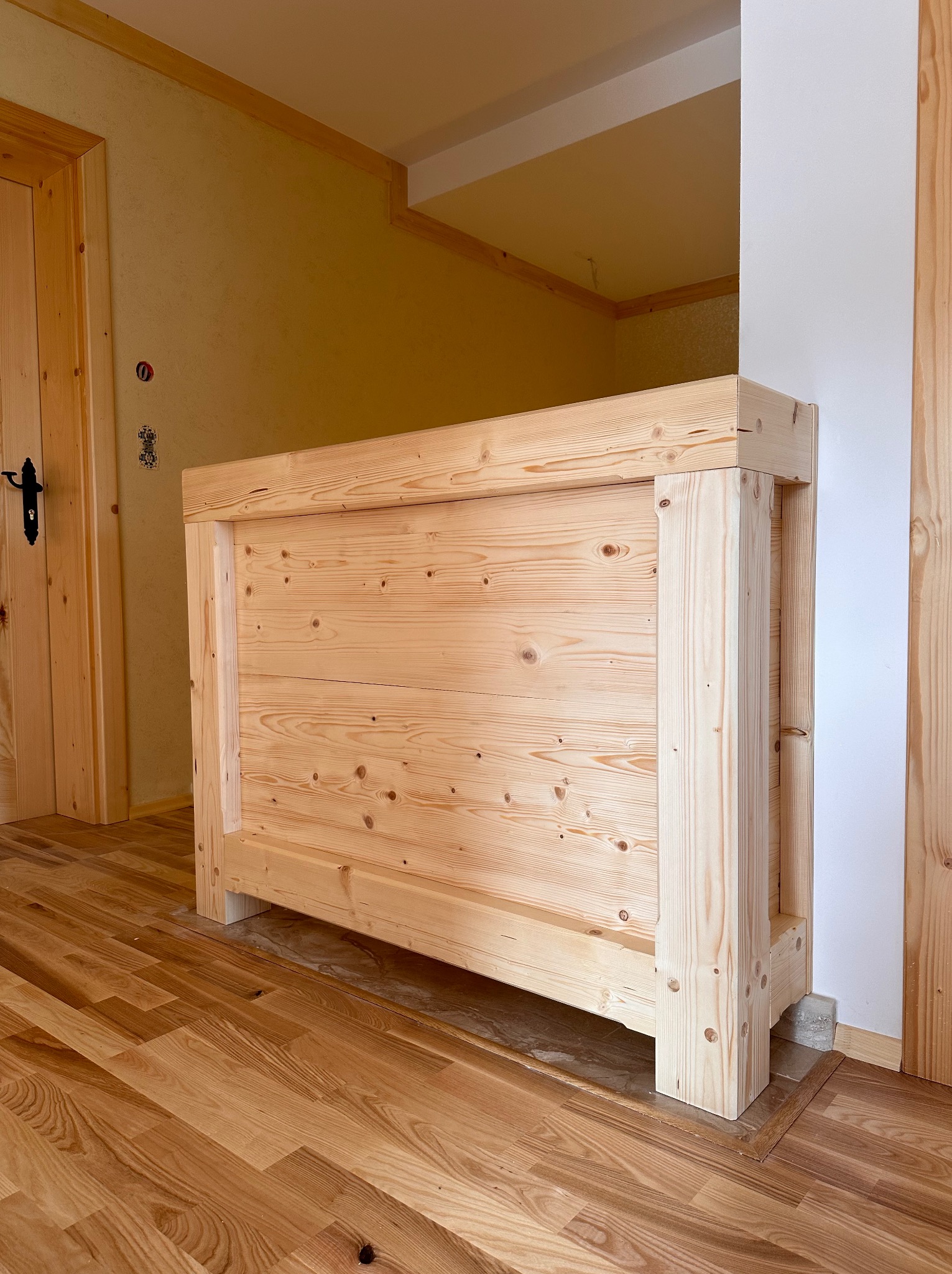 Drewniana zabudowa grzejnika z surowego drewna, widoczna na tle drewnianej podłogi i ściany. Prosta konstrukcja z desek i belek, w naturalnym odcieniu drewna.