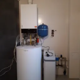 Biały, wiszący piec grzewczy Viessmann z podłączonymi rurami, zbiornikiem wyr&oacute;wnawczym i zmiękczaczem wody w jasnym pomieszczeniu z ciemną ścianą. Widoczne elementy instalacji i podłączenia.