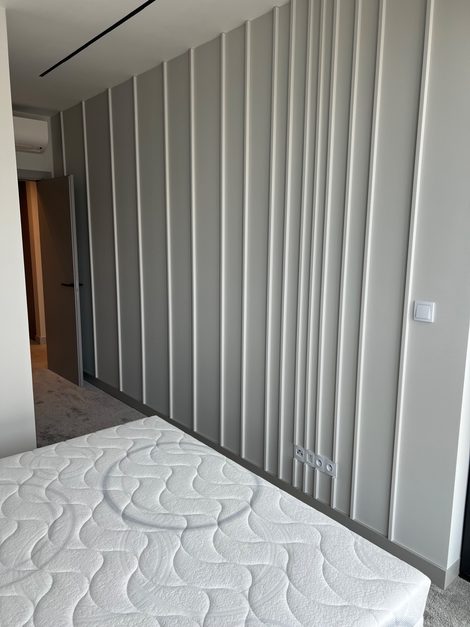 Szara ściana z pionowymi panelami dekoracyjnymi w sypialni, widoczne gniazdka elektryczne i fragment białego materaca.