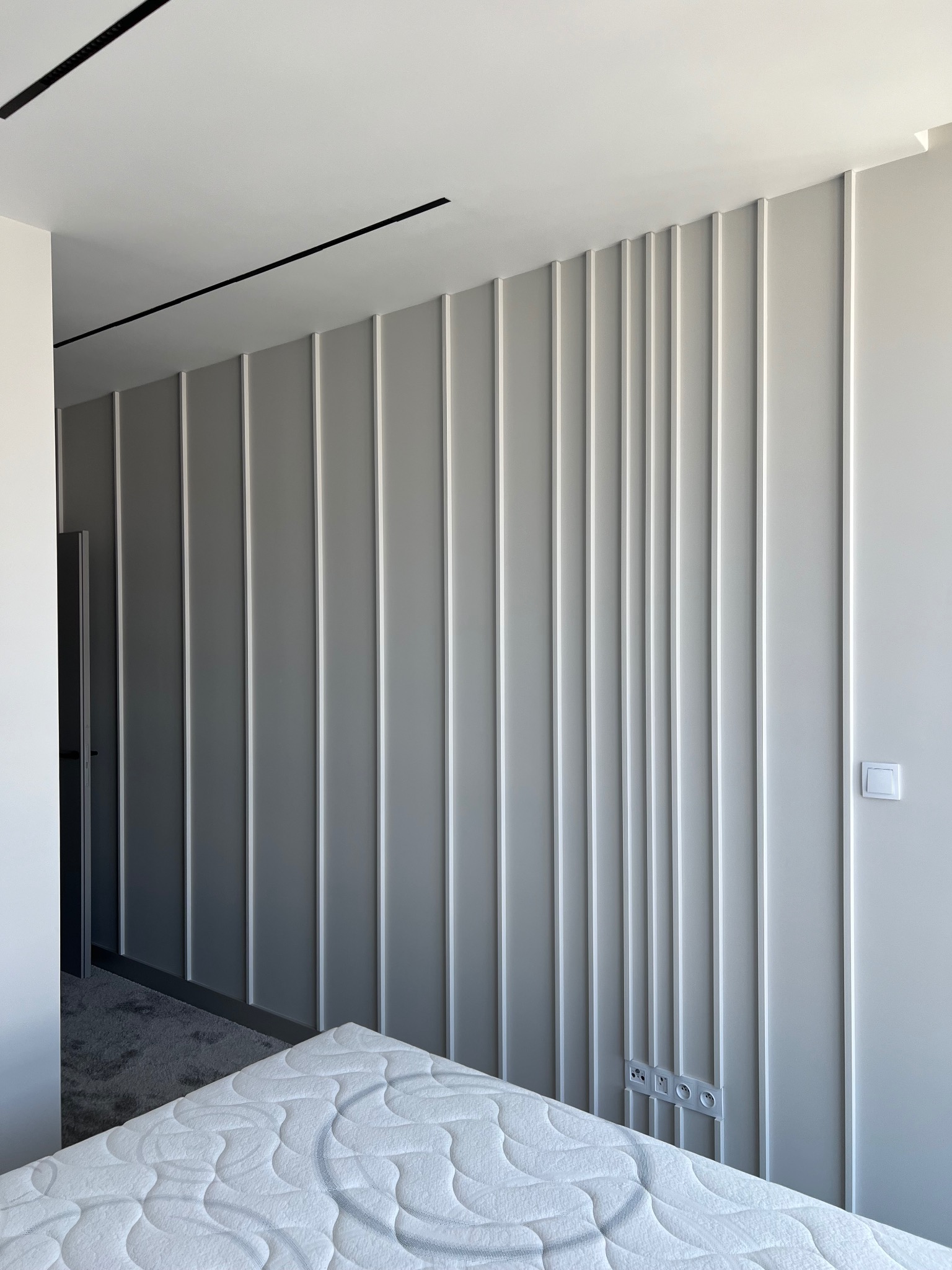 Nowoczesna sypialnia z szarą ścianą dekoracyjną z pionowymi listwami, widoczne gniazdka elektryczne, włącznik światła i fragment materaca.