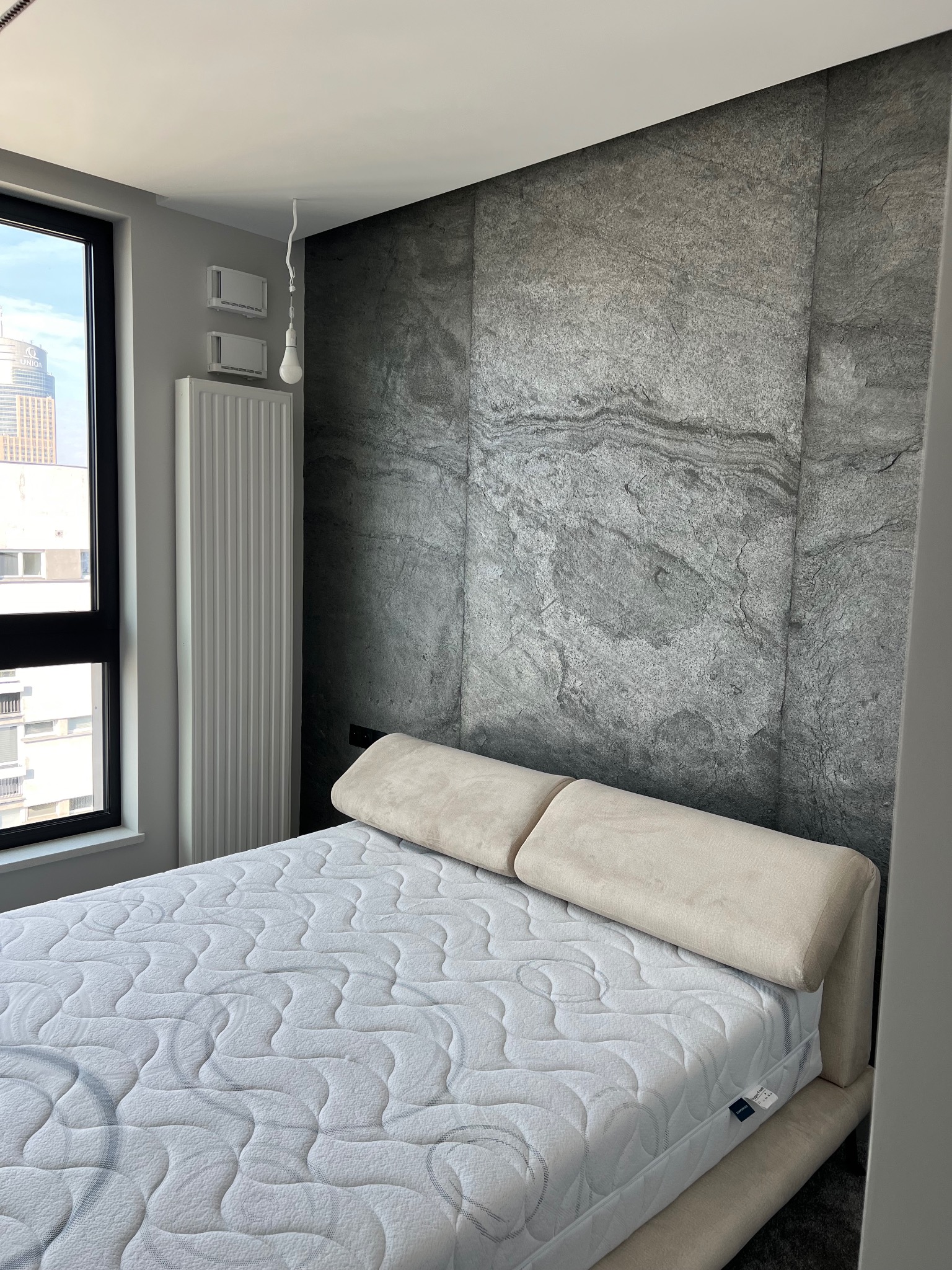 Sypialnia z łóżkiem na tle ściany wykończonej panelami z szarego kamienia imitującego marmur, widok na miasto przez okno.