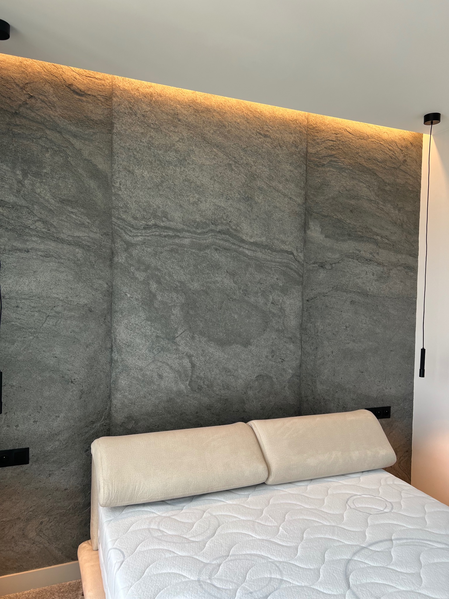 Ściana za łóżkiem wykończona szarymi płytami o strukturze kamienia, podświetlona od góry ciepłym światłem, z minimalistyczną lampą wiszącą po prawej stronie.