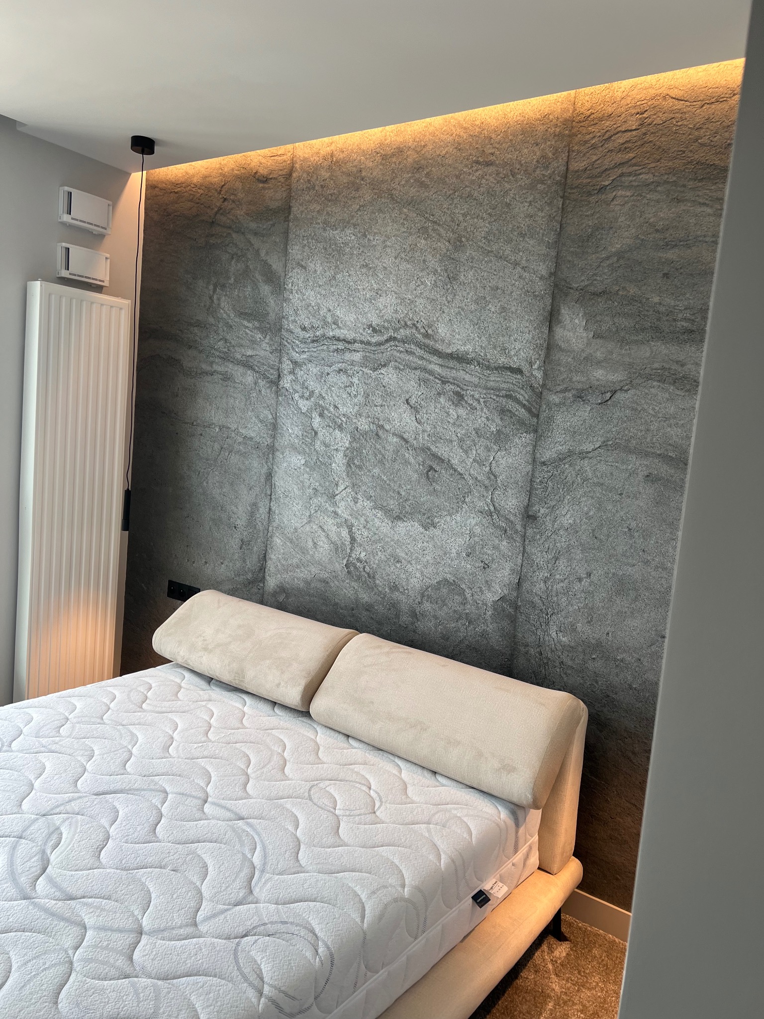 Sypialnia z łóżkiem o beżowej ramie i materacem ze wzorem, na tle ściany wykończonej panelami imitującymi szary kamień, oświetlonymi od góry delikatnym światłem.