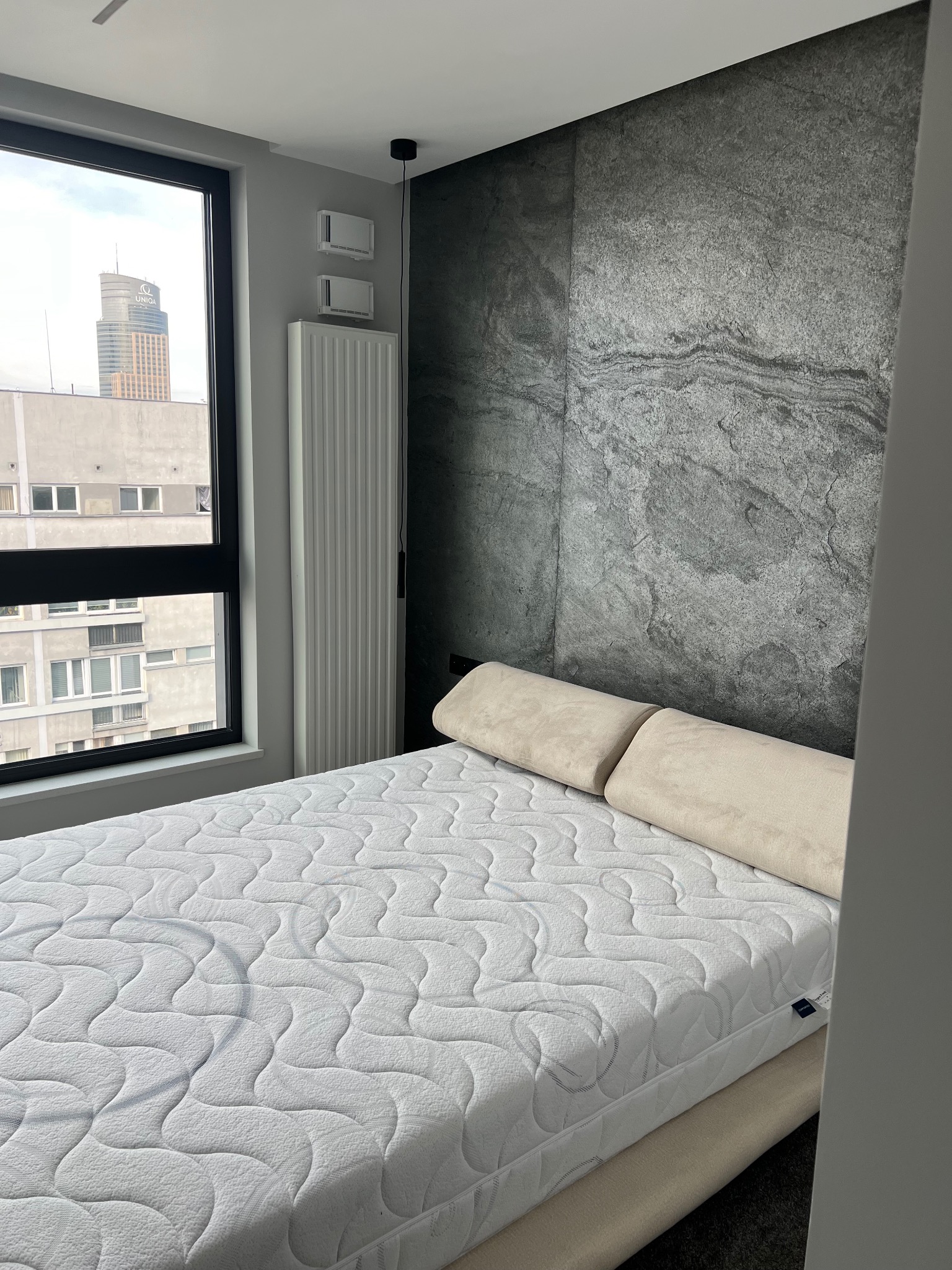 Sypialnia w nowoczesnym stylu z łóżkiem z białym materacem i dwoma poduszkami, ściana za łóżkiem wykończona szarym kamieniem dekoracyjnym, w tle widok na miasto przez okno.
