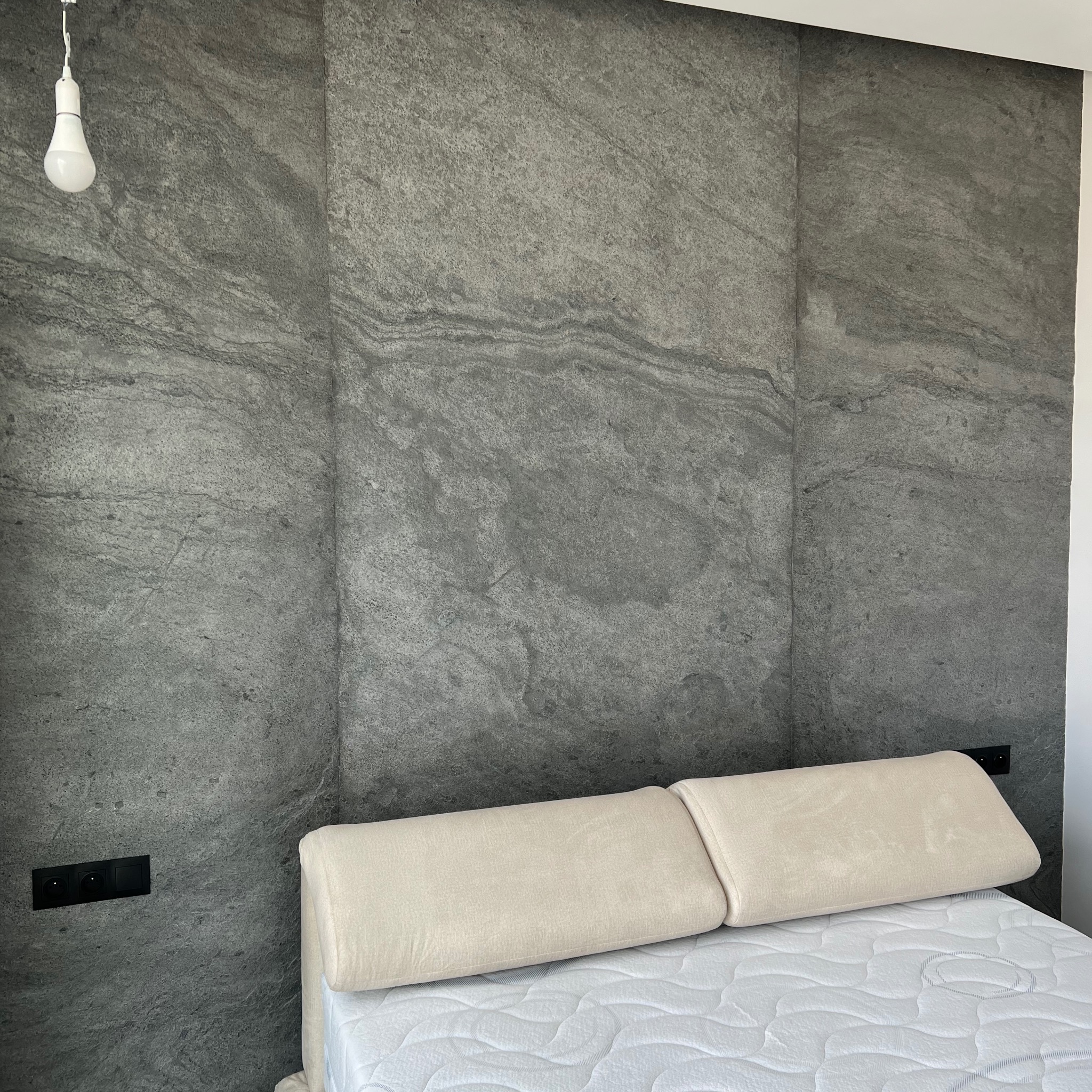 Ściana za łóżkiem wyłożona szarymi płytami imitującymi kamień, z widocznymi czarnymi gniazdkami elektrycznymi i wiszącą żarówką na kablu.