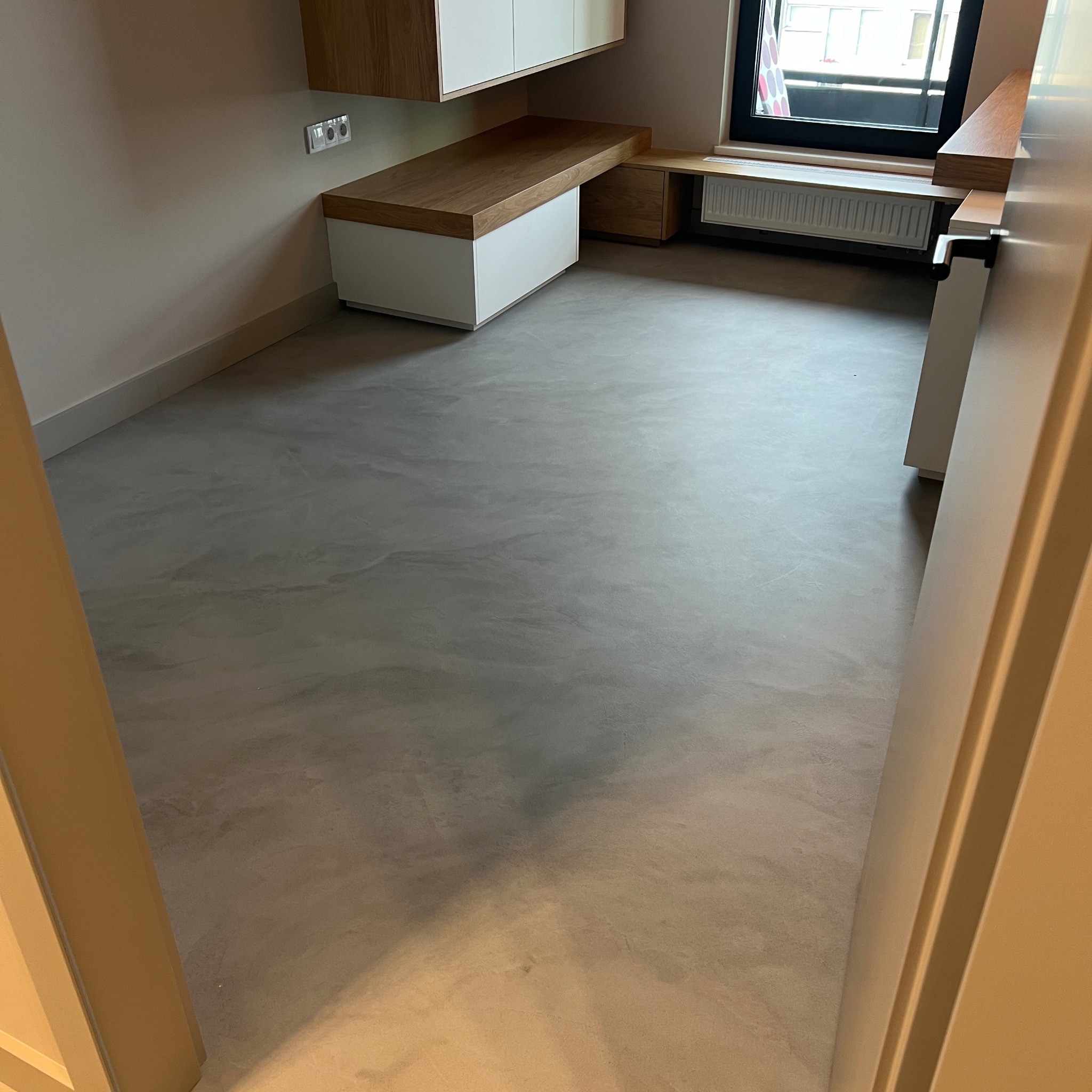 Mikrocementowa posadzka w jasnym pomieszczeniu z zabudową meblową z drewna i bieli, widok z progu drzwi.
