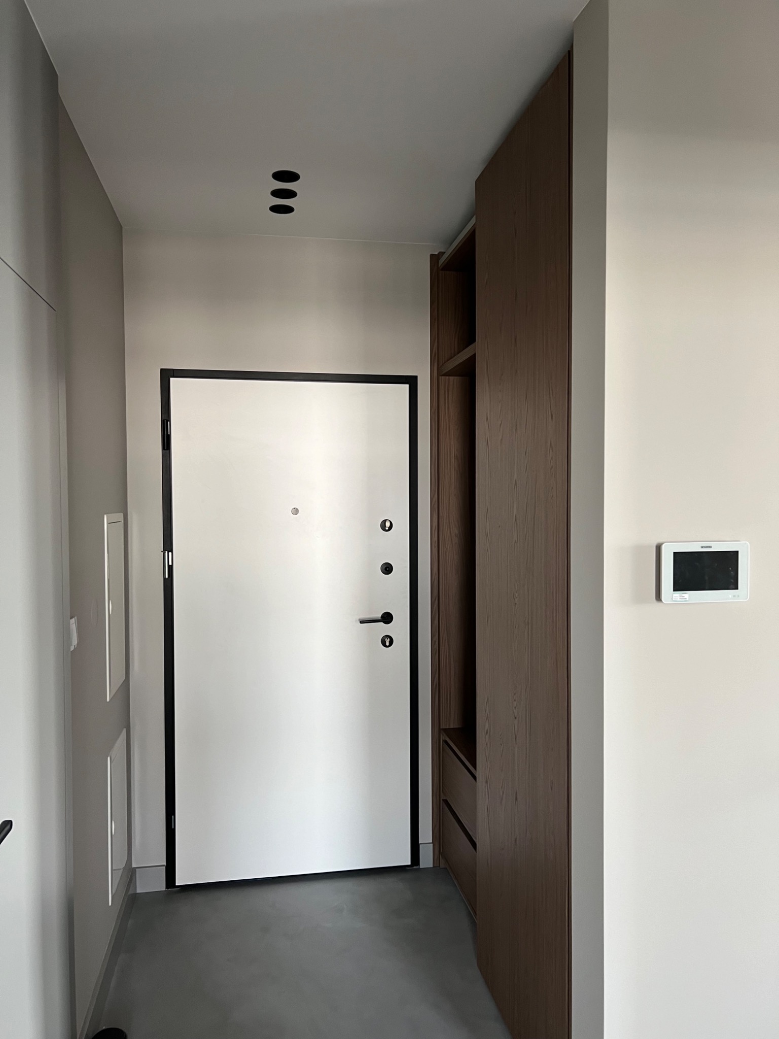 Nowoczesny przedpokój z białą, antywłamaniową  drzwi wejściowymi z czarną ramą, wbudowaną szafą z ciemnego drewna i panelem sterowania na ścianie.