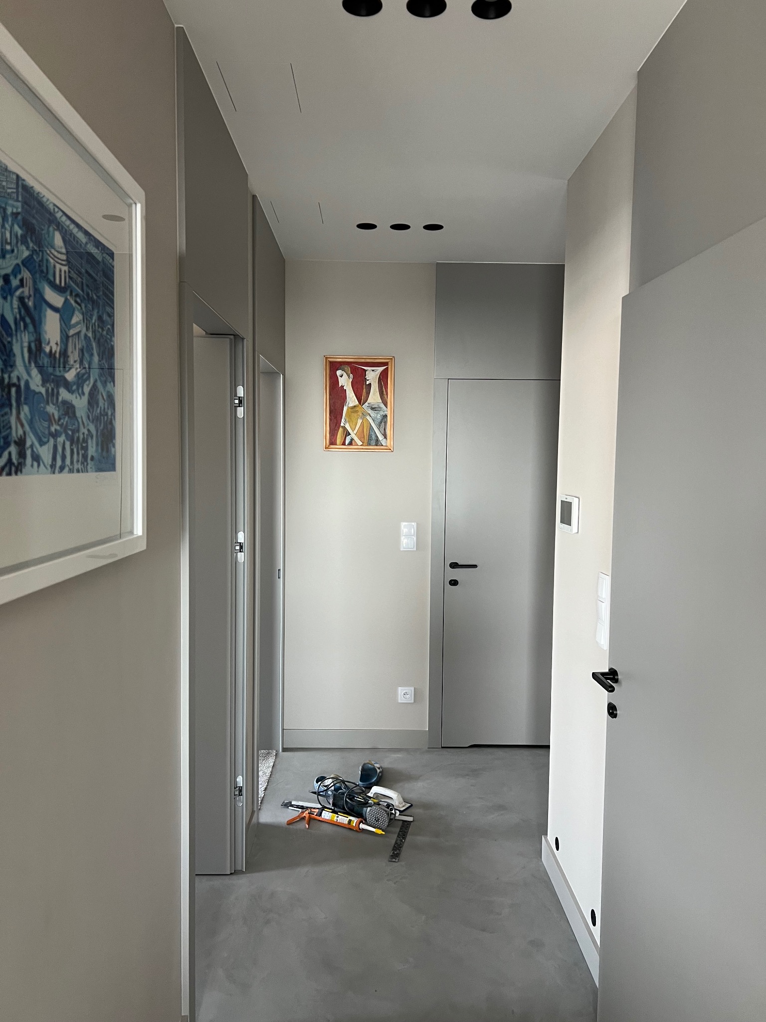 Korytarz w odcieniach szarości z betonową podłogą, oświetleniem punktowym i dwoma obrazami na ścianach, z narzędziami do wykończenia podłogi leżącymi na posadzce.