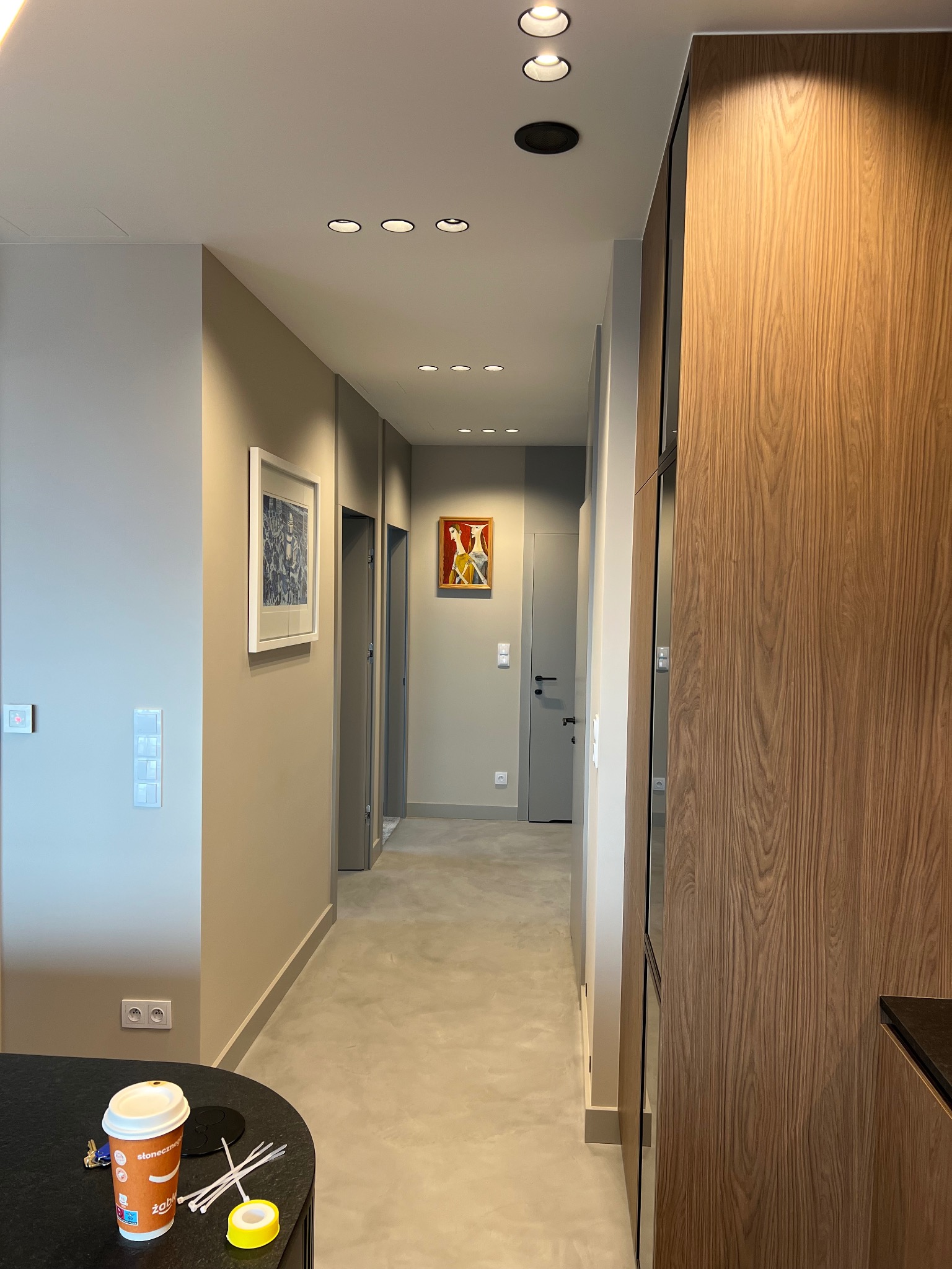 Nowoczesny korytarz z szarą podłogą, beżowymi ścianami, drewnianą zabudową i oświetleniem punktowym, z widocznymi detalami jak obrazy na ścianach i elementy instalacji elektrycznej.