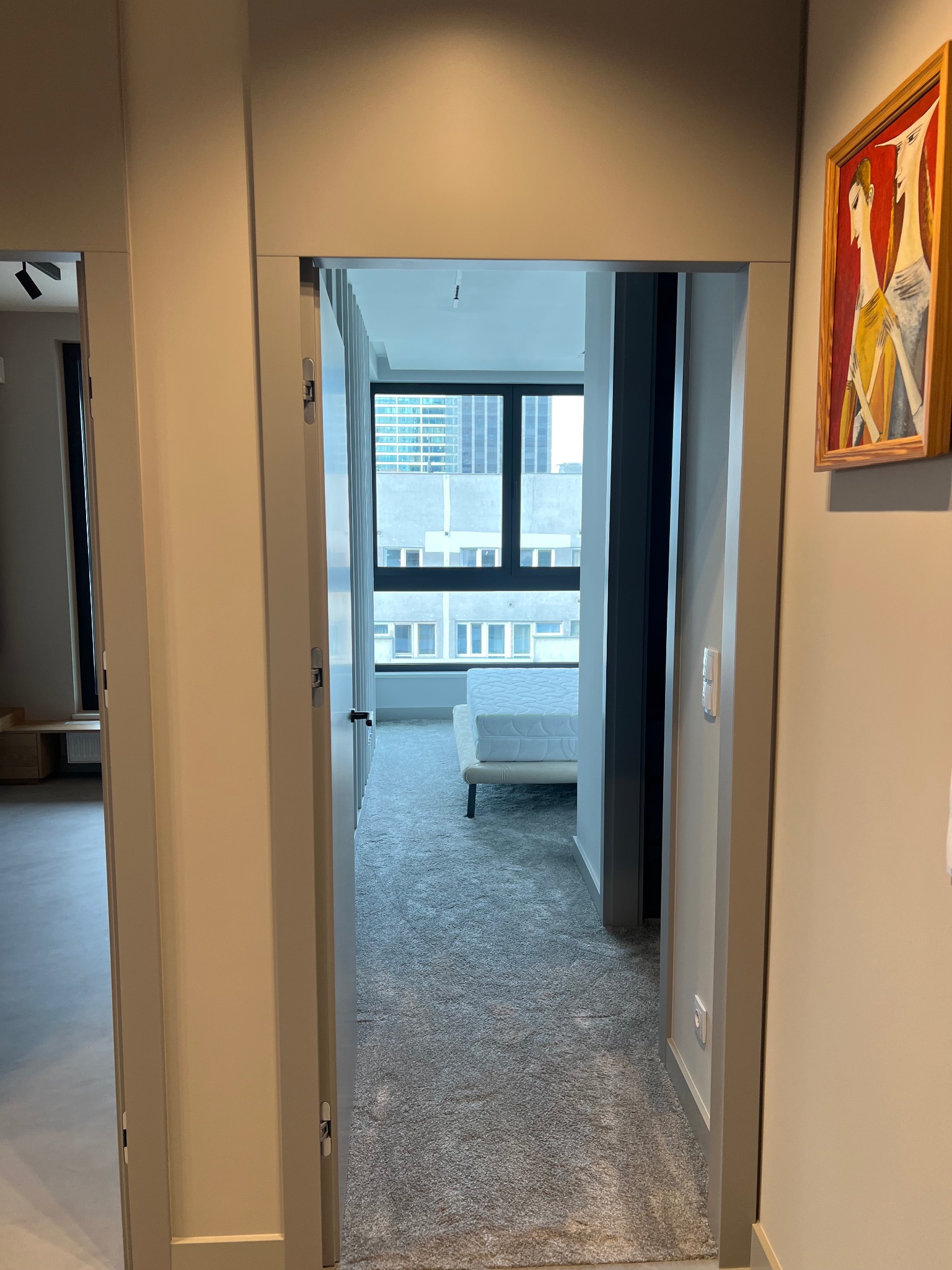 Wnętrze mieszkania z widokiem na sypialnię przez otwarte drzwi, na ścianie wisi obraz w drewnianej ramie.