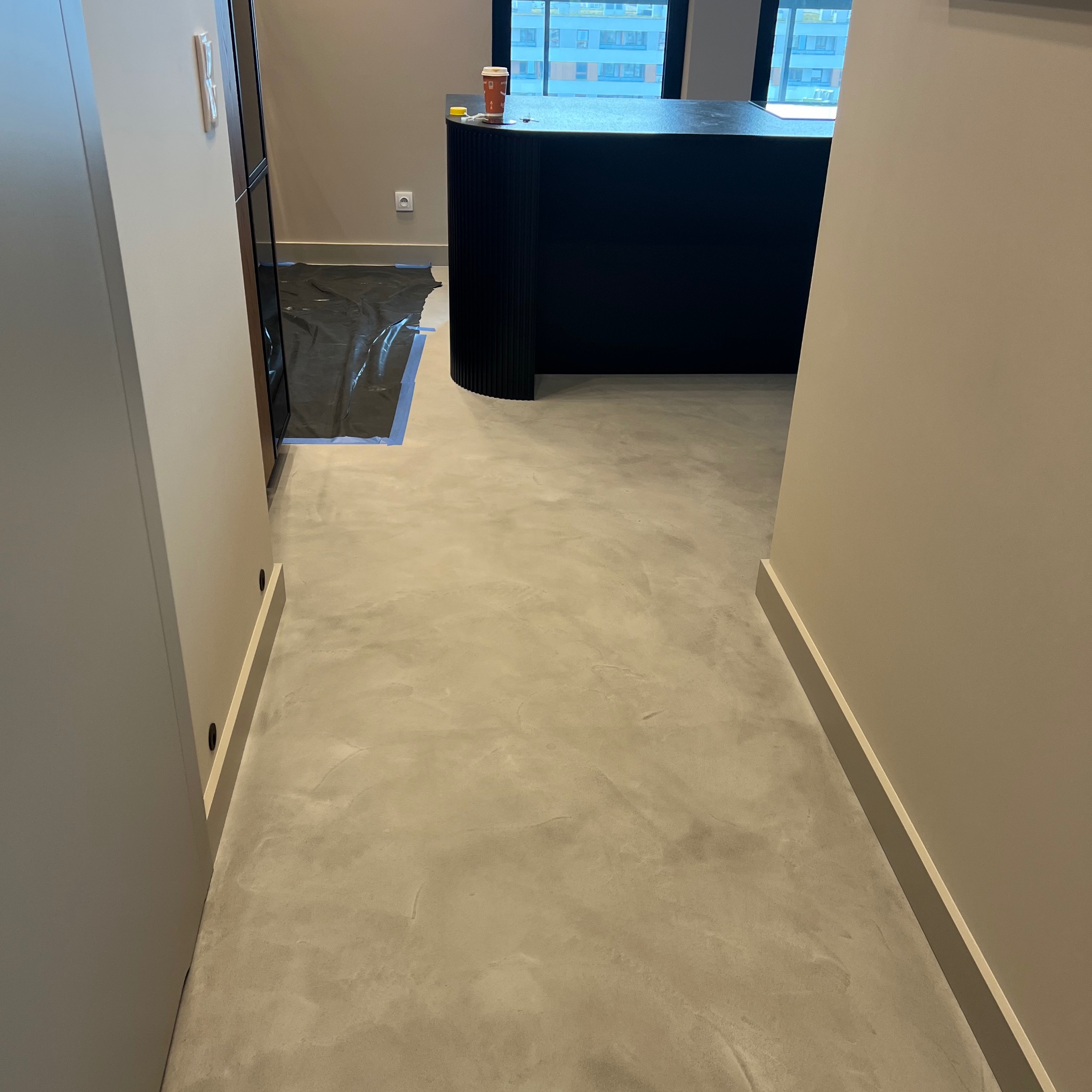 Wypoziomowana, szara posadzka w korytarzu, zabezpieczona folią przy czarnym barze z żłobieniami, widok z perspektywy wejścia.