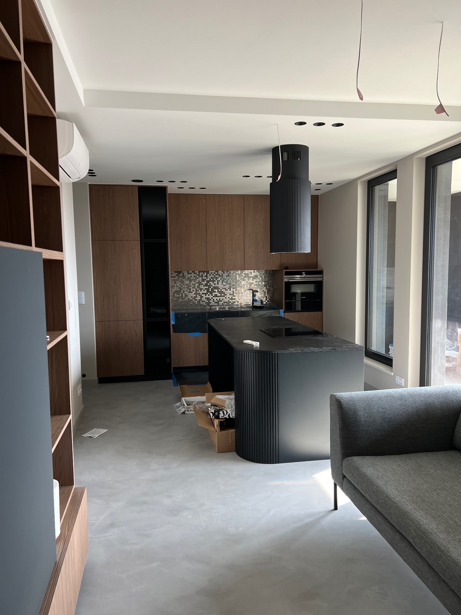 Wnętrze kuchni w trakcie wykańczania: drewniane szafki, czarna wyspa z ryflowanym wykończeniem, mozaika nad blatem, widoczne kartony i elementy montażowe na podłodze.