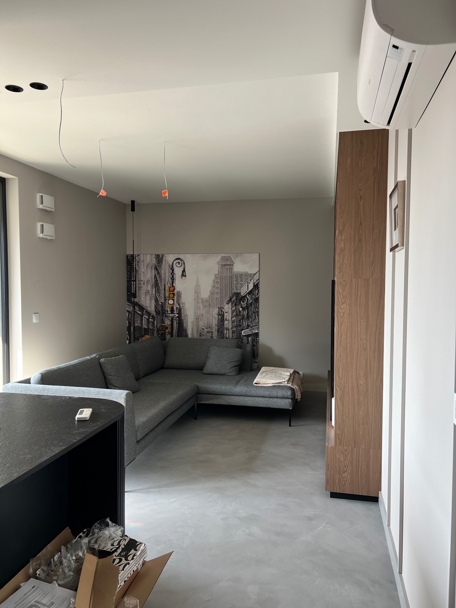 Nowoczesny salon z szarą sofą narożną, betonową podłogą i obrazem miejskiego krajobrazu na ścianie. Widoczne niedokończone instalacje oświetleniowe na suficie i klimatyzator.
