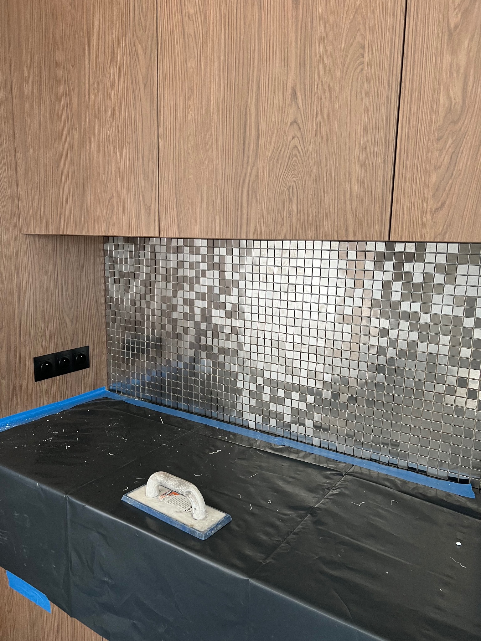 Montaż mozaiki srebrno-szarej na ścianie, widoczna paca do nakładania kleju, zabezpieczona powierzchnia robocza folią i taśmą malarską, drewniane szafki wiszące powyżej.